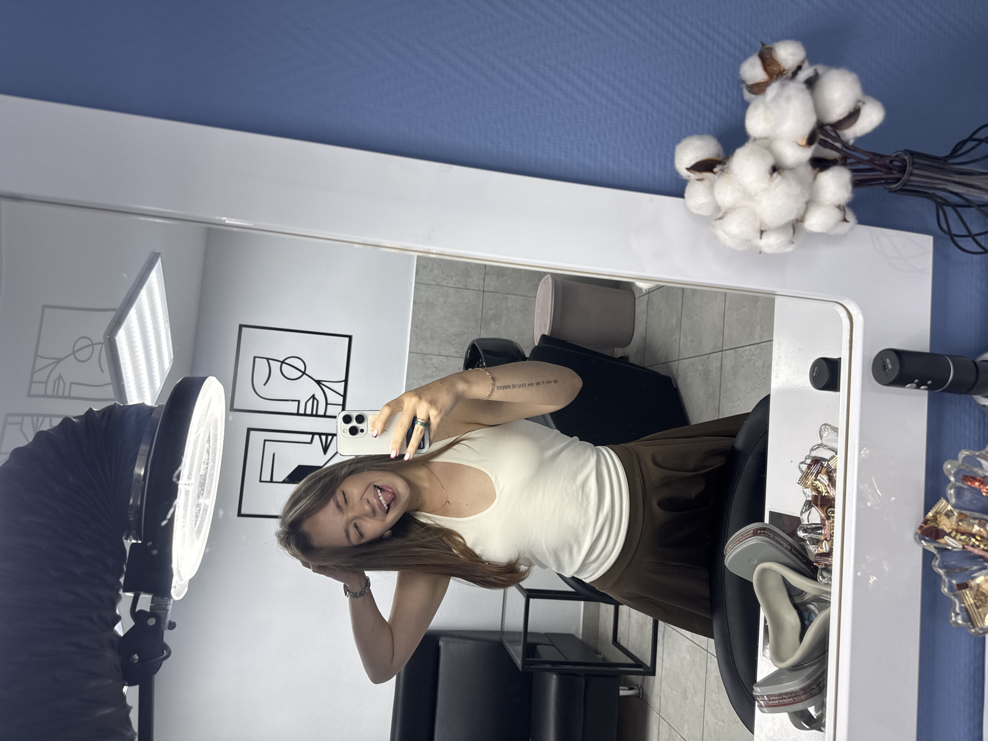 A young woman with long brown hair is taking a selfie in a brightly lit room with blue walls. She is wearing a white halter top and a brown skirt. She is sticking her tongue out and winking at the camera. In the background, there is a black leather chair, a sink, and a trash can. There are also two framed abstract drawings on the wall. On a shelf in front of the mirror, there is a pair of gray goggles and some wrapped candies. To the right of the mirror, there is a vase with cotton balls. The location is a beauty salon in the city of Chaikovsky, Russia.