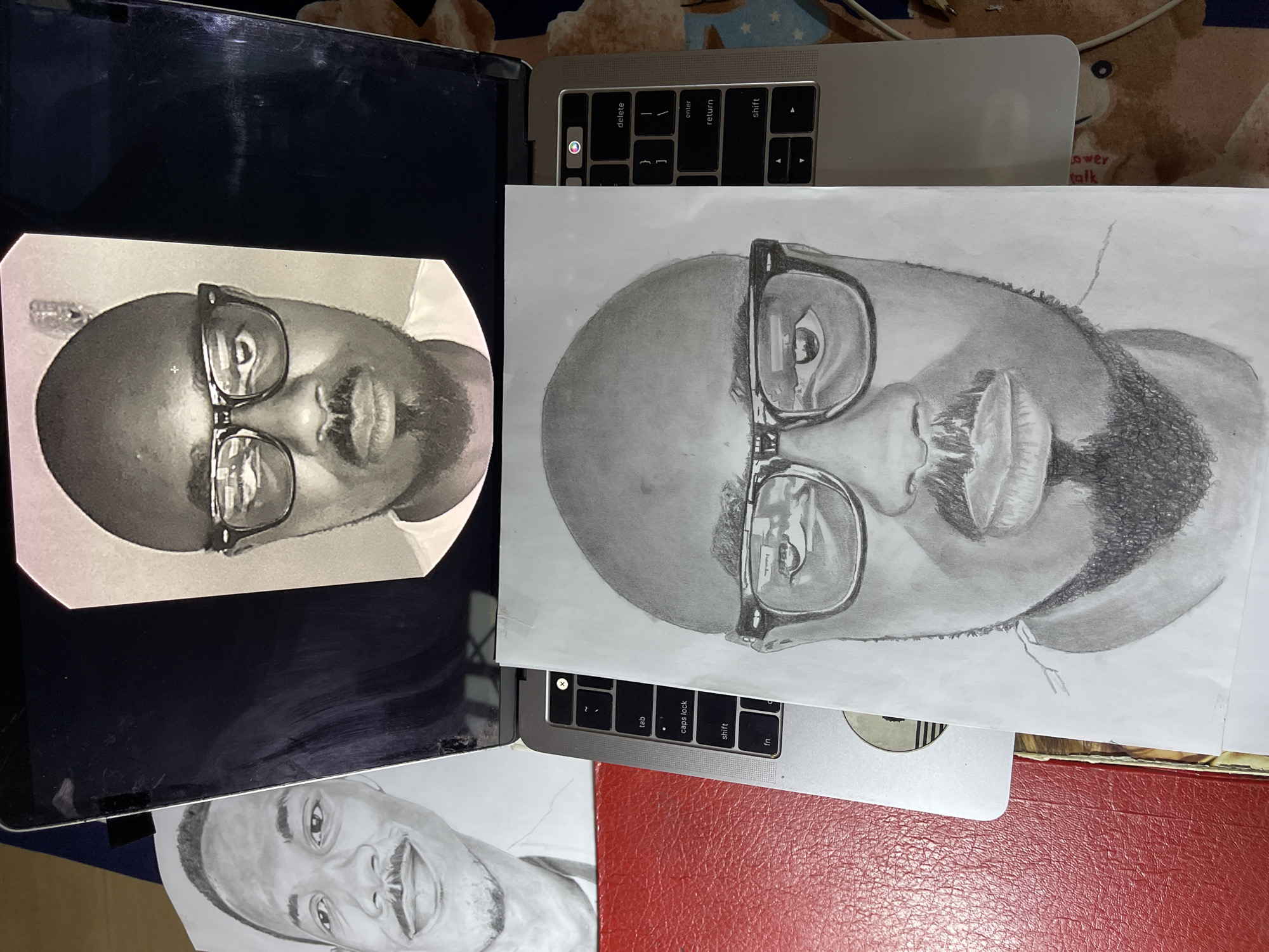 The scene depicts a collection of objects on a flat surface with patterned fabric visible in the background. Two open laptops are present, each displaying a portrait of a man with glasses. A physical drawing of the same man also sits between the laptops, slightly overlapping. One laptop screen displays the portrait digitally, while the other contains the physical drawing directly on top. The background hints at an indoor setting, potentially in a private residence given the presence of the laptop and personal items. This image provides no information that would directly pinpoint it to Jos, Nigeria.