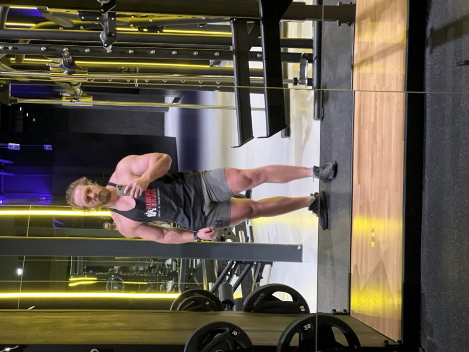 A muscular man stands in a gym, reflected in a mirror, holding a phone up to take a picture. He wears a black tank top and grey shorts. Behind him is gym equipment: racks, cables, and weights. Yellow lighting illuminates the scene. The mirror's reflection shows more of the gym's interior.