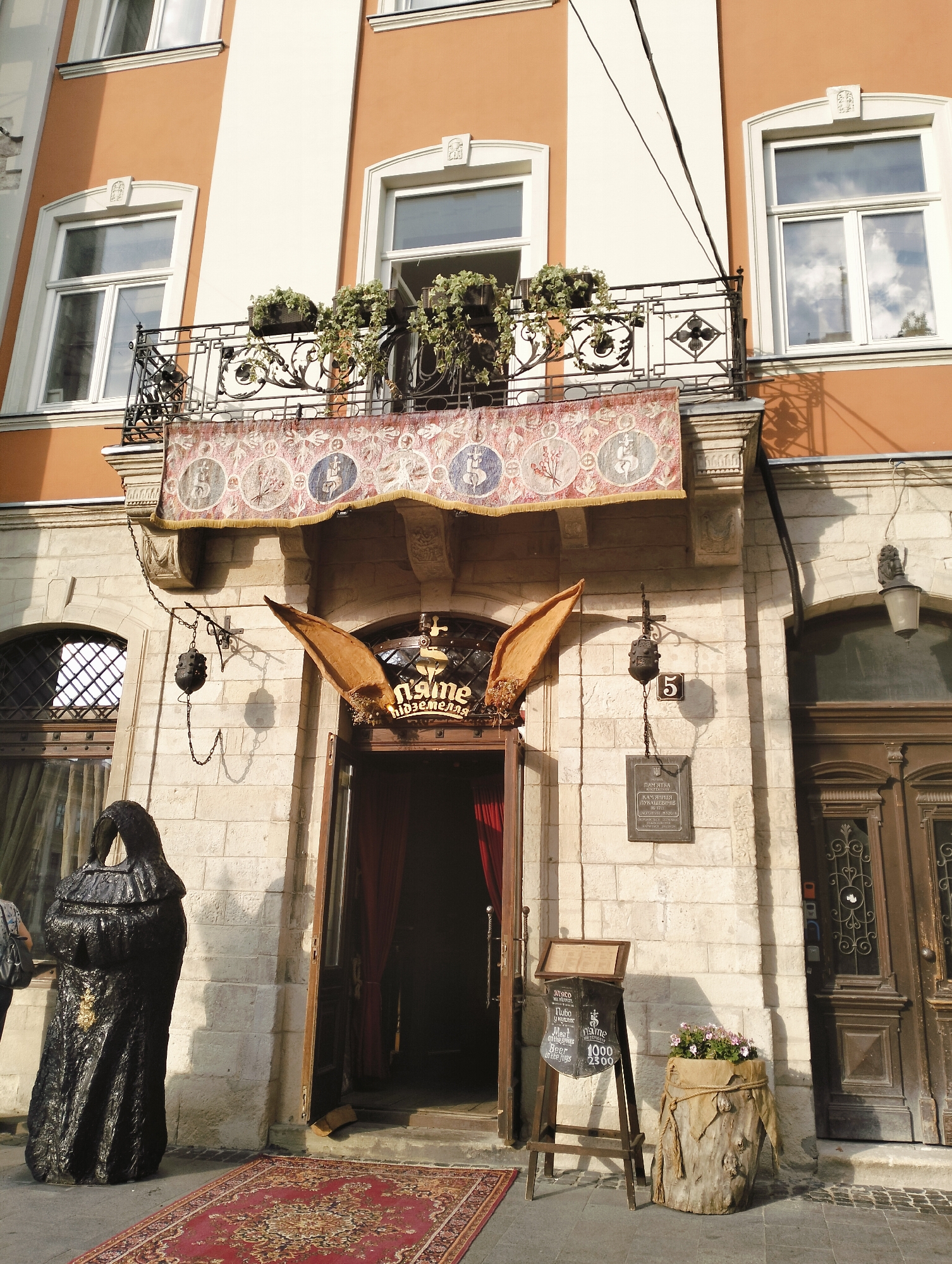 This is a front-facing, eye-level, medium shot of the entrance to the "Kryivka" restaurant, a themed establishment in Lviv, Ukraine, known for its World War II Ukrainian Insurgent Army (UPA) theme. The building's facade is constructed of light-colored stone blocks. Above the entrance, a balcony with an ornate iron railing features potted green plants. A decorative fabric banner with circular emblems hangs below the balcony.
The entrance itself is an open doorway framed by wooden doors, with red velvet curtains visible inside. Above the doorway, large, stylized wooden wings are mounted, and a sign with Cyrillic script and what appears to be a crown is also present. To the left of the entrance, a tall, black statue resembling a cloaked figure stands on a red patterned rug. To the right of the entrance, a street sign with the number "5" is mounted on the wall. A plaque with Ukrainian text is also affixed to the wall near the number "5". Further to the right, a closed wooden door with decorative metalwork is visible.
In front of the restaurant, a wooden easel displays a chalkboard menu with prices and offerings, including "Wine," "Auto – Auto," and "Beer." Next to the easel, a large planter made of rough wood, tied with rope, holds pink flowers. The ground in front of the entrance is paved with stone, and a red oriental rug lies on the pavement leading up to the doorway. The upper portion of the image shows parts of the building's upper floors, including windows with white frames and orange walls. Sunlight casts shadows on the facade.