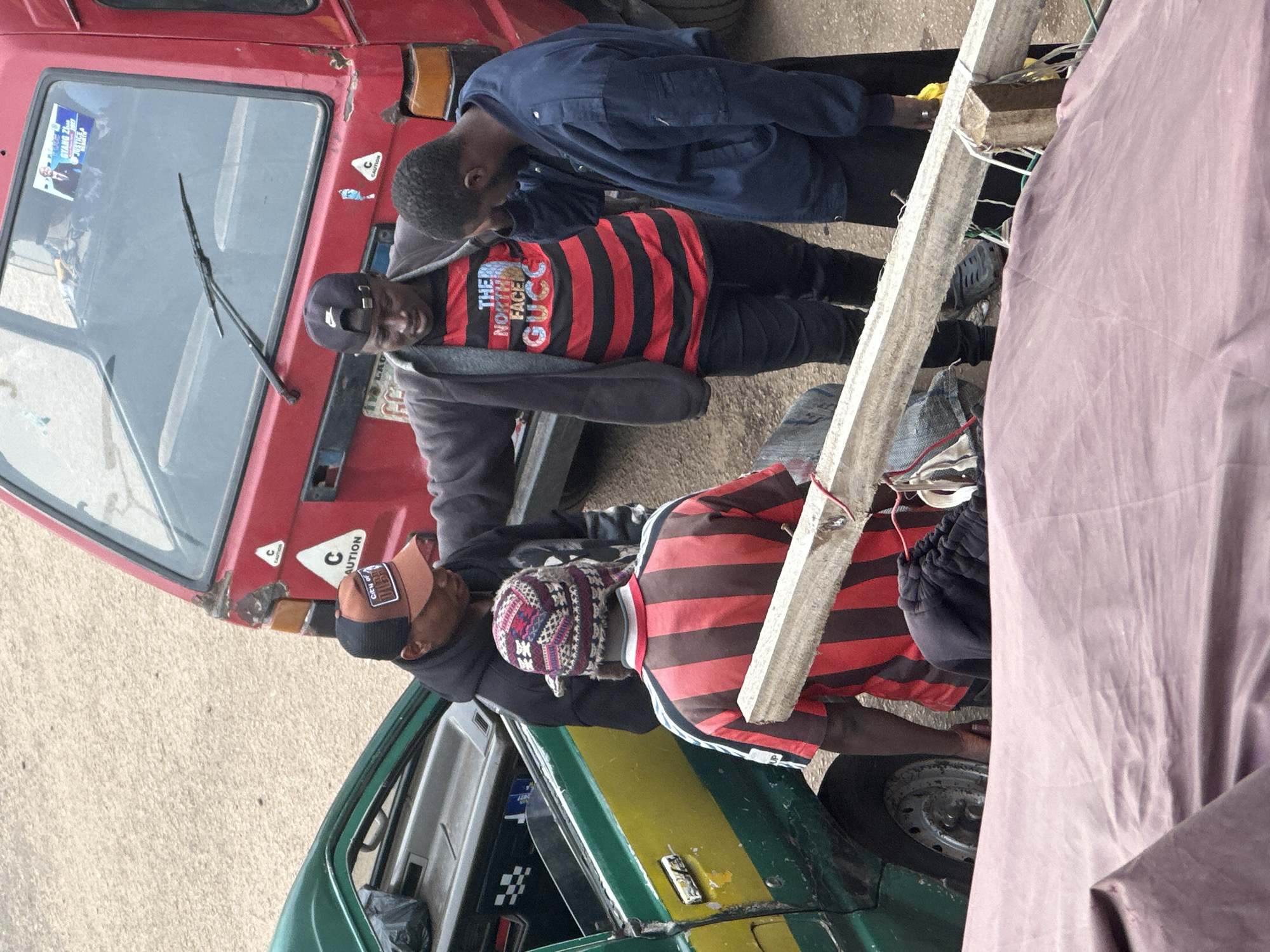 In Bukuru, Nigeria, a scene unfolds with four men in close proximity, appearing to be engaged in a conversation or transaction. Two men stand near the open door of a green and yellow taxi, one wearing a black hoodie and a brown cap, the other a patterned beanie and a red and black striped shirt. They are positioned between the taxi and a red vehicle, possibly a truck. A third man, dressed in a gray jacket over a red and black striped shirt with "The North Face Gucci" text, stands facing them, his hand extended. A fourth man, in a dark blue jacket, stands slightly apart from the group, his head bowed. The ground is paved, and a wooden beam secured with rope lies in the foreground, partially obscured by a pink cloth. The overall atmosphere suggests a busy urban environment.