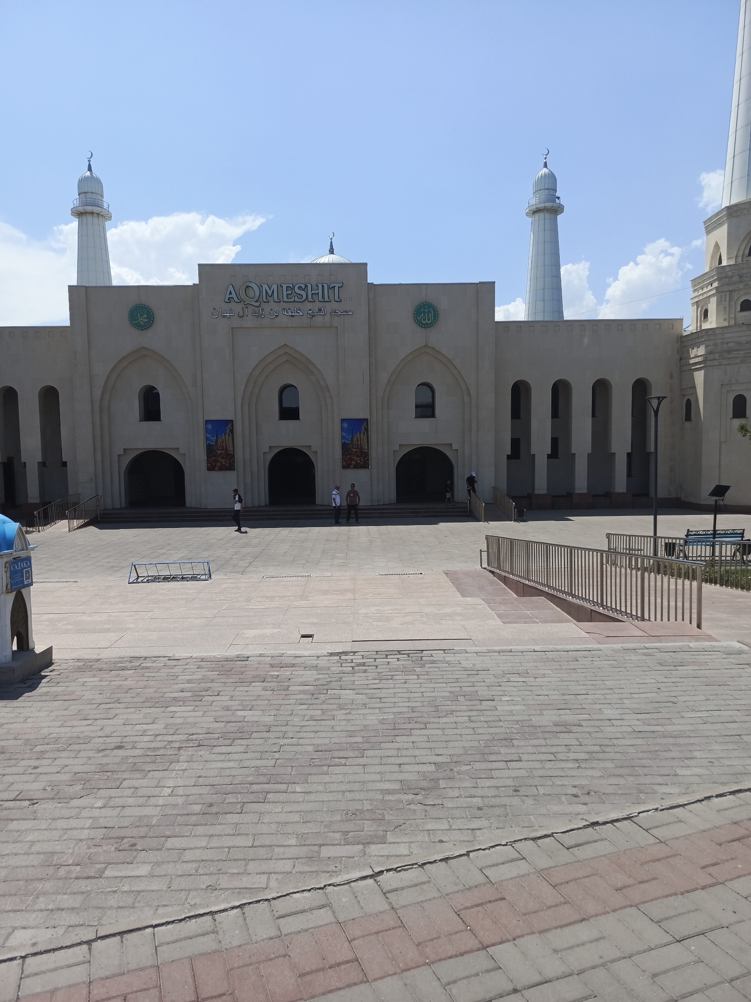 The image shows the exterior of the Aqmeshit Mosque in Shymkent, Kazakhstan. The mosque features a light-colored facade with arched entrances and tall minarets topped with crescent moons. Arabic calligraphy is visible above the main entrance, as well as a circular emblem with Arabic script. Two large banners depicting architectural scenes hang on either side of the central arched entrance. The mosque is situated on a wide, paved plaza with brickwork. Several people are present in the plaza, including a few individuals walking or standing near the mosque entrance, and others further in the distance. A metal bench and a bicycle rack are visible on the plaza. The sky is blue with scattered white clouds.