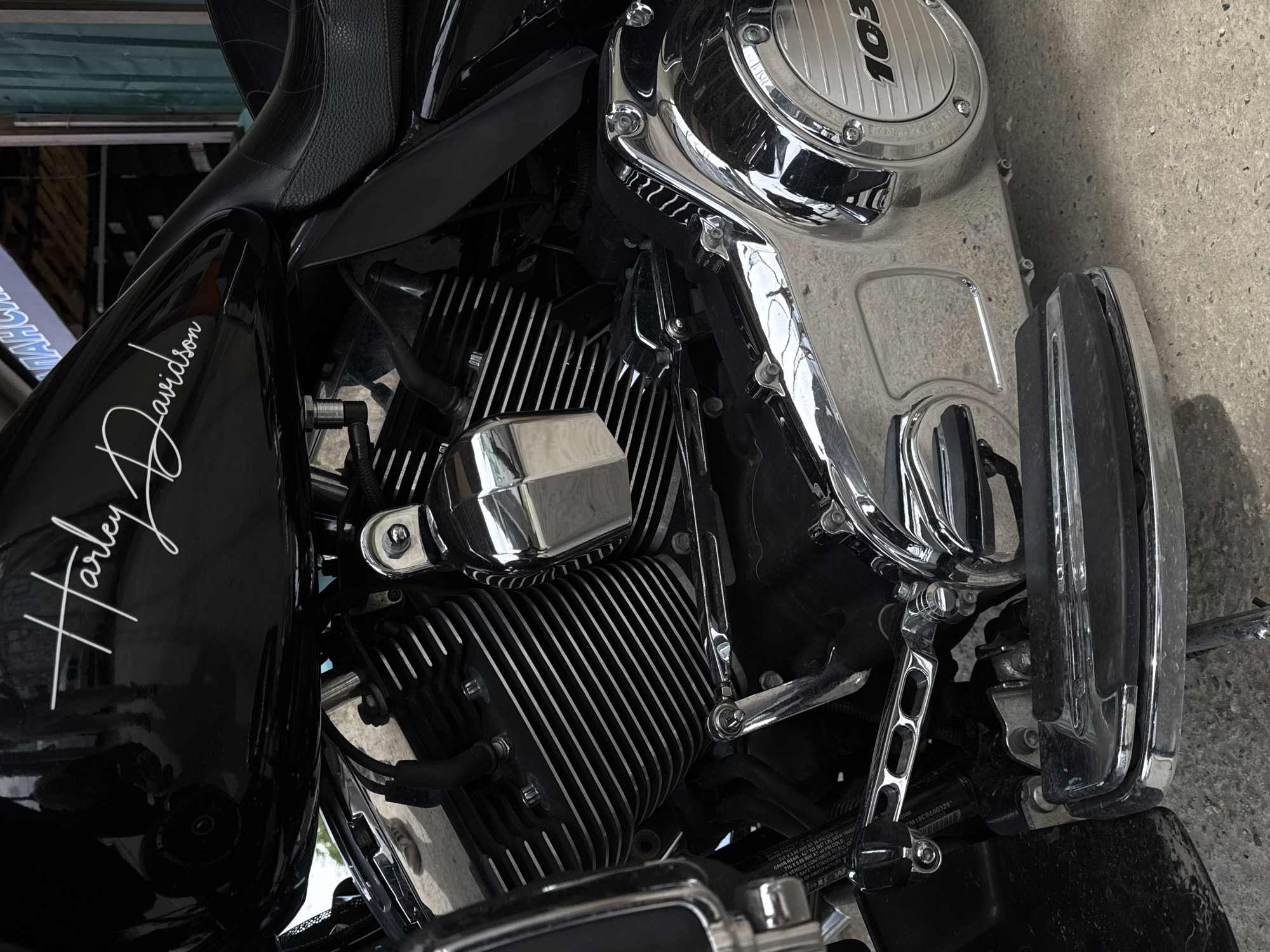 The image shows a close-up of a Harley Davidson motorcycle engine. The engine is black and chrome, with the "Harley Davidson" logo visible on the side. The engine size "103" is on the top of the engine. The engine has a cylinder head with cooling fins. The motorcycle is on a concrete surface. There are some words printed on the frame of the motorcycle. The words "RIM AT PSI COLD", "FORMS TO A", "SAFETY STANDARDS", "OF MANUFACTURE", "GAWR REAR-421 KG (927 LR)", "IS VEHICLE CONTO", "CM18CB600329". There is no evidence to support the location as Krasnodar, Russia.