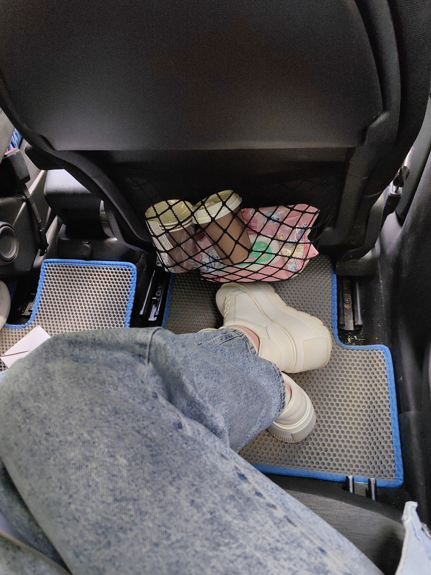 The image shows the interior of a vehicle, likely a car. A person is seated with their legs and feet visible. They are wearing blue jeans and white platform sneakers. The person's left leg is extended forward, resting on a gray car mat with a blue trim. In the background, a mesh organizer is attached to the back of a car seat. This organizer contains two disposable coffee cups and a pink bag with a pattern of stars and a cartoon character. The scene appears to be indoors or in a covered area, as indicated by the limited natural light and the presence of car interior elements. There is no information to suggest the location is in the city of Сваткаўскі сельскі Савет, Belarus.