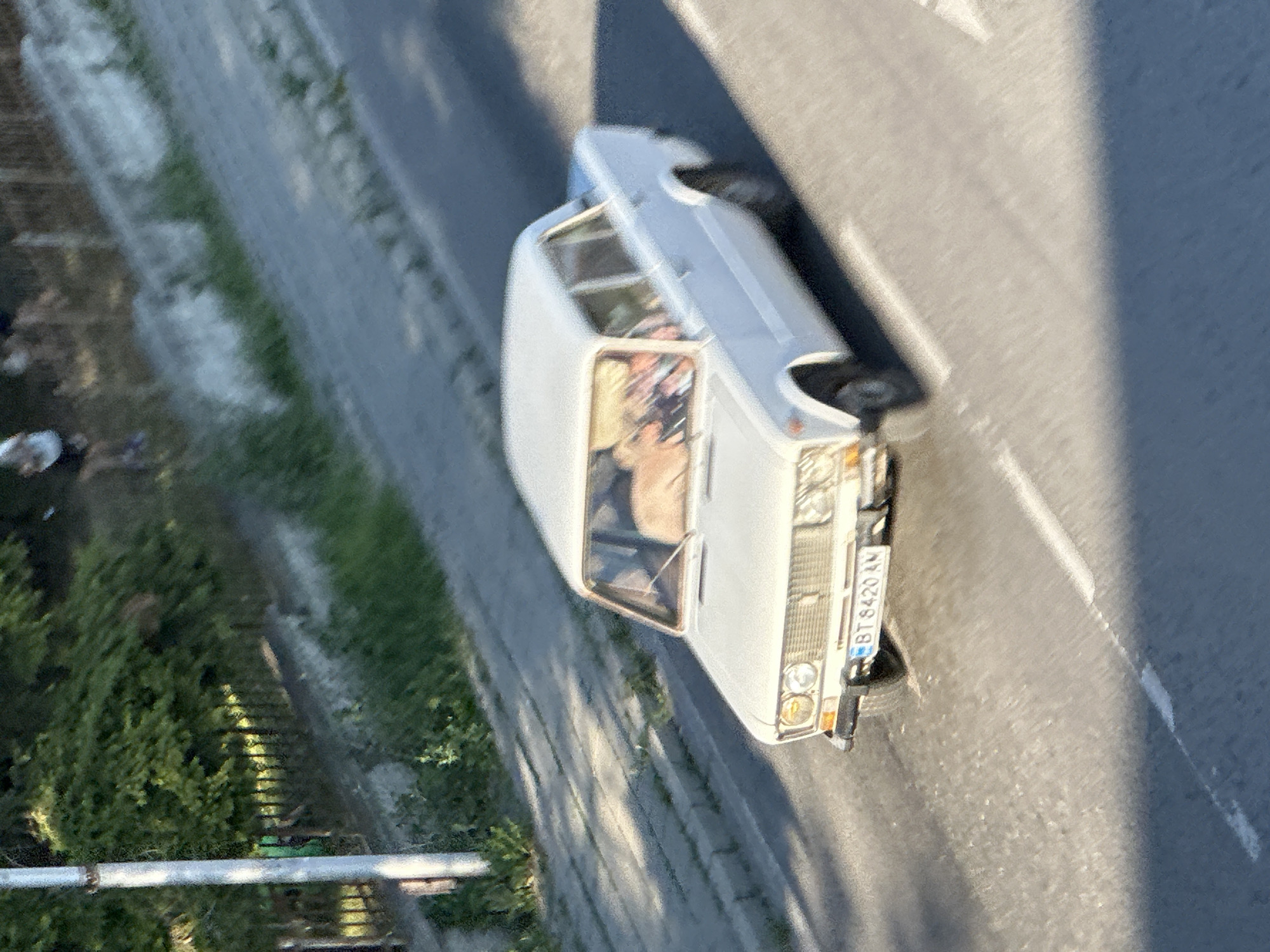 The image captures a white sedan parked on a paved road, likely in an urban or semi-urban environment given the presence of a sidewalk and adjacent greenery. The car's license plate is visible, reading "BT 8420 AY". Sunlight casts shadows, suggesting it is daytime, possibly late afternoon or early morning due to the length of the shadows. The setting appears to be a street with trees and vegetation lining one side. The car is positioned at an angle, allowing a view of its front and side. The windows of the car are slightly tinted, and one can discern the shapes of people inside. The overall impression is of a still moment, capturing a vehicle in its environment. The scene is set in Veliko Tarnovo, Bulgaria.