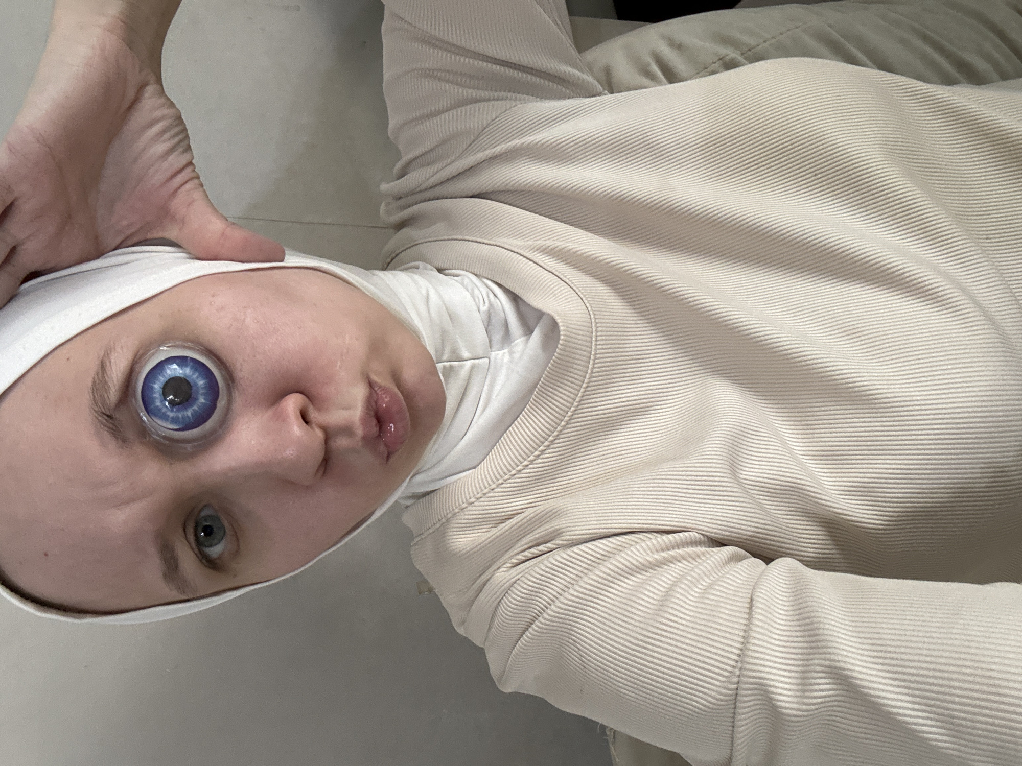 A person is pictured from a low-angle perspective, wearing a white head covering and a cream-colored, ribbed long-sleeve shirt. The person is lying on their side, with their head tilted back. Their right eye appears to have an unusual, large, blue prosthetic eye. The person is puckering their lips slightly. A hand is raised above their head. The background consists of light gray and cream-colored surfaces, possibly a floor and a couch. There is no information in the provided image or prompt that indicates the location is the city of Chaikovsky, Russia.