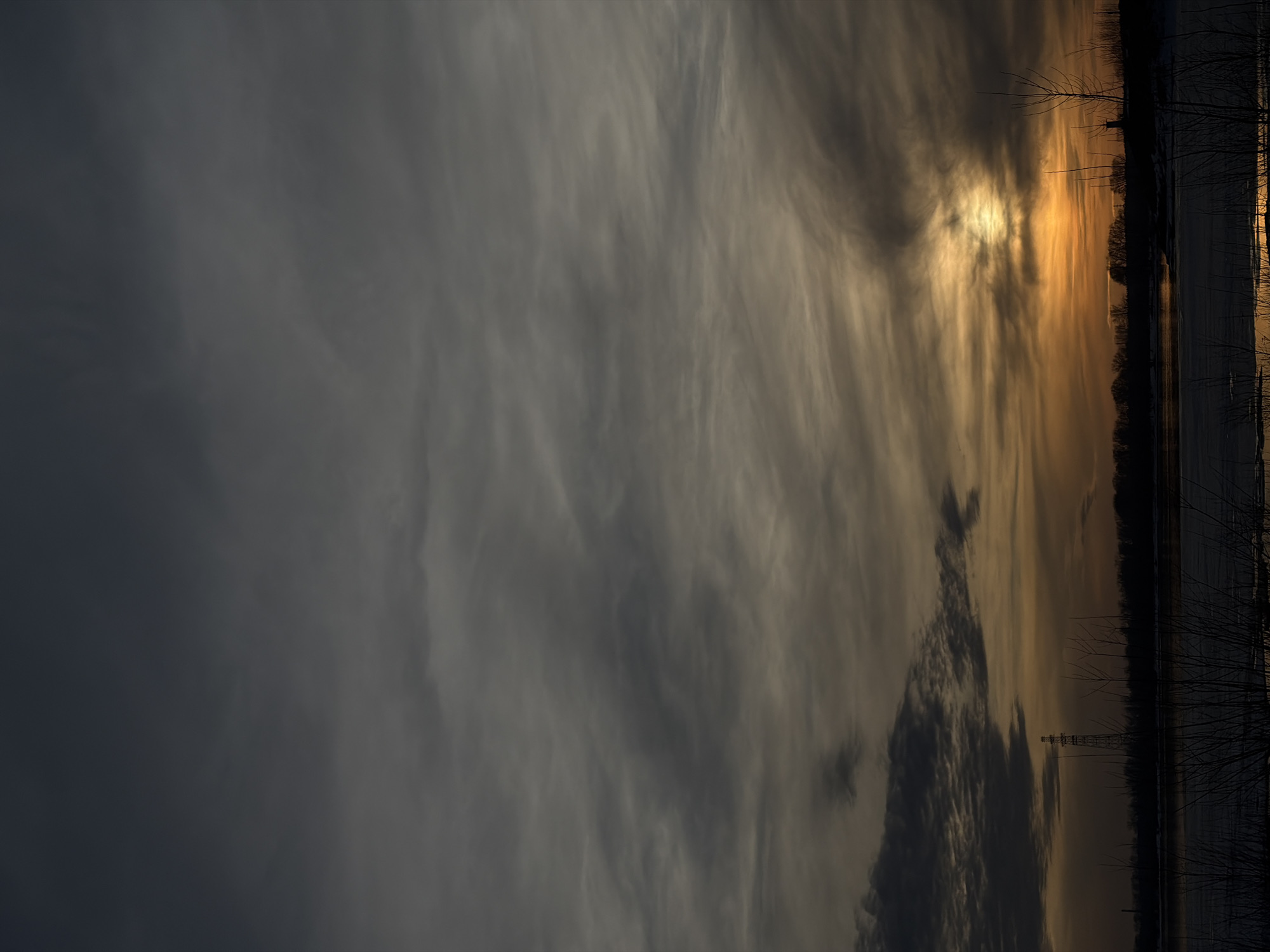 The image captures a twilight landscape in Chaikovsky, Russia. The sky, occupying the majority of the frame, is overcast with layers of dark grey and lighter grey clouds. Towards the lower right, the setting sun is partially obscured by clouds, creating a concentrated patch of amber and yellow light that extends horizontally along the horizon, gradually fading into muted orange and diffused gold tones. This illumination reflects on a body of water in the midground, which appears partially frozen with segments of open, reflective water. The horizon line consists of a dark, silhouetted strip of what appears to be a treeline, likely deciduous, with indistinct distant structures. On the left side, a tall, lattice-style electrical transmission tower stands silhouetted against the illuminated sky, emerging from the tree line. The immediate foreground displays dark, indistinct bare branches and sparse vegetation. No human figures are present.