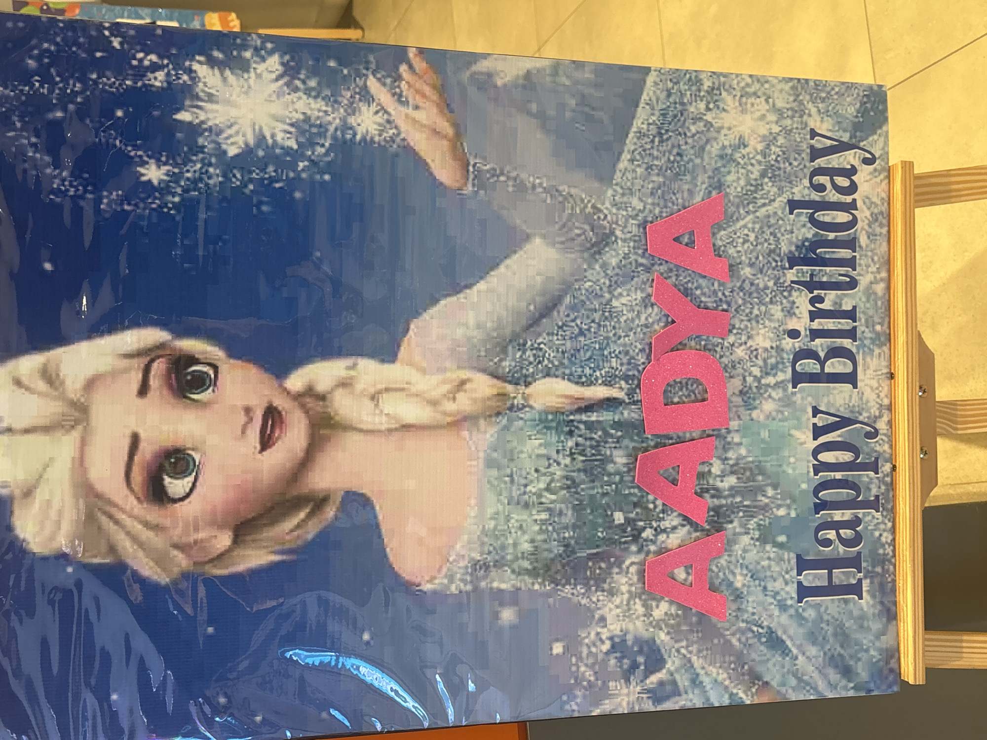 A digital artwork of Elsa from Frozen is displayed on a wooden easel. Elsa is depicted from the chest up, with blonde hair in a braid, wearing a light blue dress with a snowflake pattern. She has large blue eyes and is smiling. Pink glitter letters spelling "AADY A" are overlaid on the image, followed by blue text that reads "Happy Birthday". The background is a blue, snowy scene with snowflakes. The easel is placed on a tiled floor.