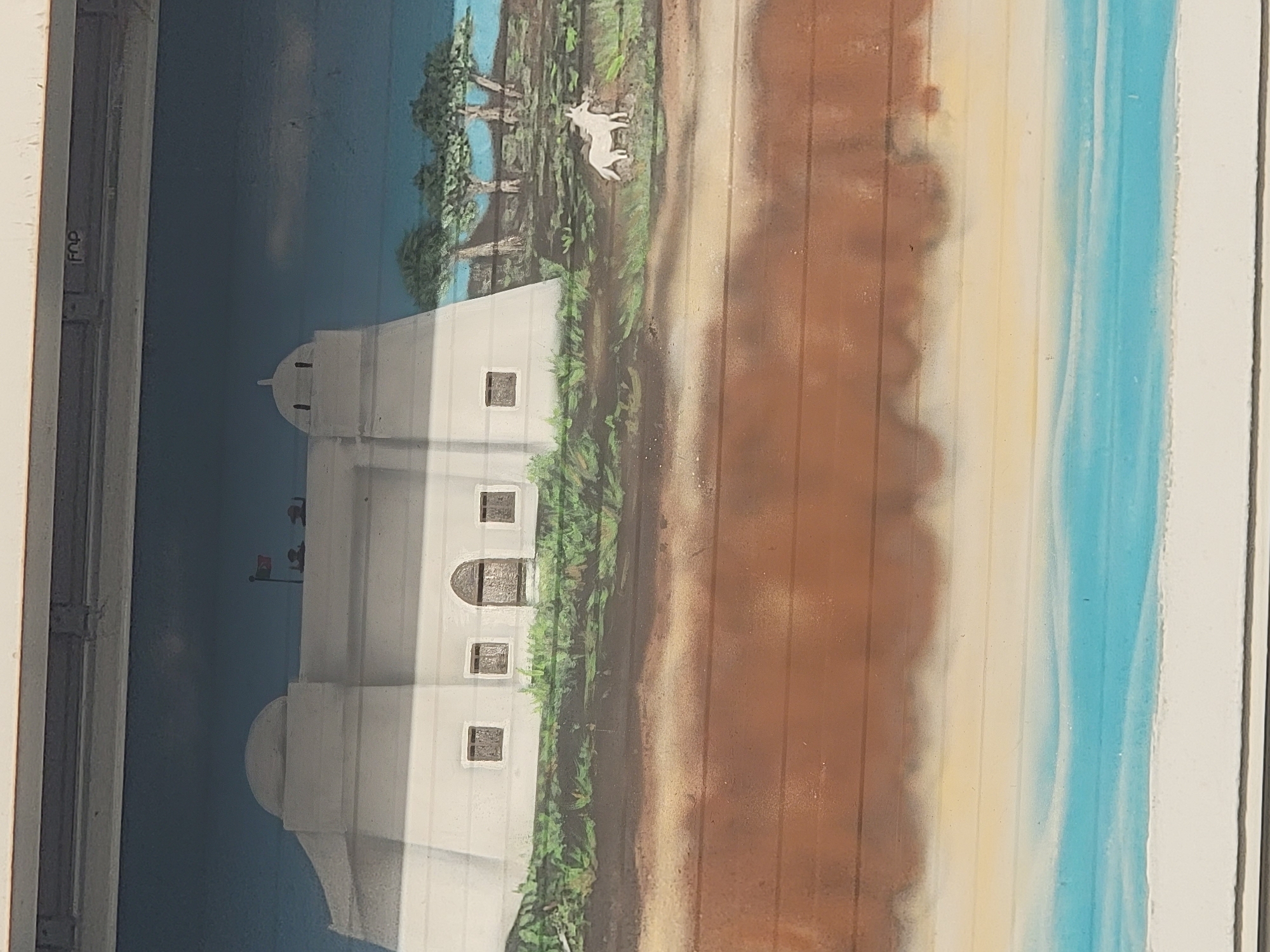 The image depicts a mural painted on a textured, horizontal surface, likely a garage door or a similar industrial facade. The mural is divided horizontally into three distinct sections. The top section features a blue sky with some clouds, and a white fortress-like building with a domed roof. The building has several small windows and a large arched doorway. A flag flies from the top of the fortress, and two figures are visible on the roof. To the right of the fortress are several trees with sparse foliage, and a small white animal, possibly a dog or a goat, stands in a grassy area.
The middle section of the mural is a sandy or earthy terrain, with subtle shading and texture suggesting ground. The bottom section of the mural depicts a stylized representation of water, with bands of blue and white, suggesting waves or the sea.
The overall style of the mural is somewhat naive and cartoonish, with bold colors and simplified shapes. The painting is located on the exterior of a building in Quarteira, Portugal.