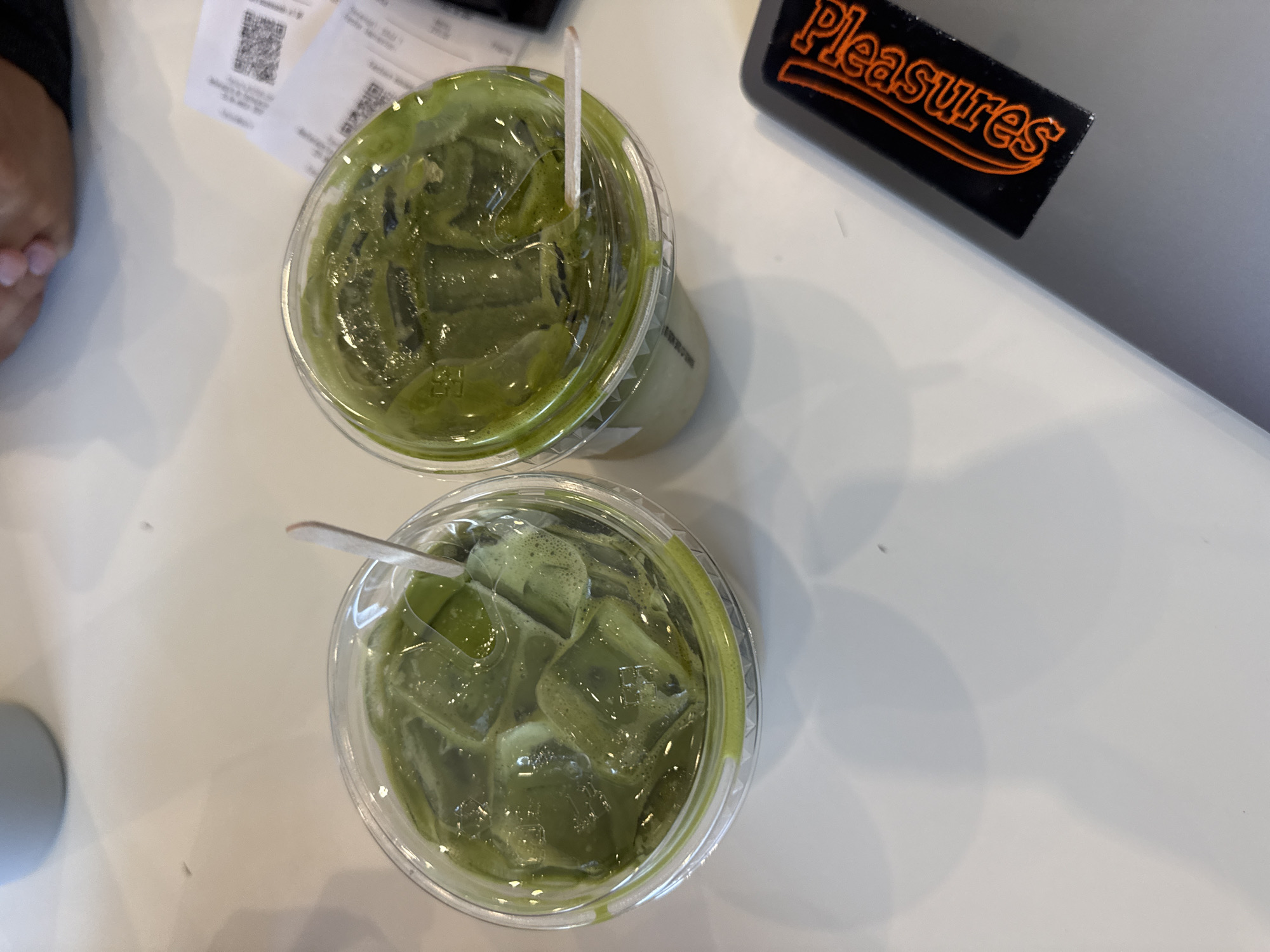 Two clear plastic cups filled with an iced green beverage, likely matcha, are prominently featured on a white table. Each cup contains ice cubes and a small wooden stirrer or spoon. In the background, to the upper right, a black object with the word "Pleasures" written in orange, stylized text is visible. Two crumpled receipts, each displaying a QR code and blurred text, are situated behind the top cup. A hand with neatly manicured nails is partially visible in the upper left corner of the frame. The overall scene suggests a casual setting, possibly a cafe or eatery, in Puebla, Mexico, where someone is enjoying cold drinks.