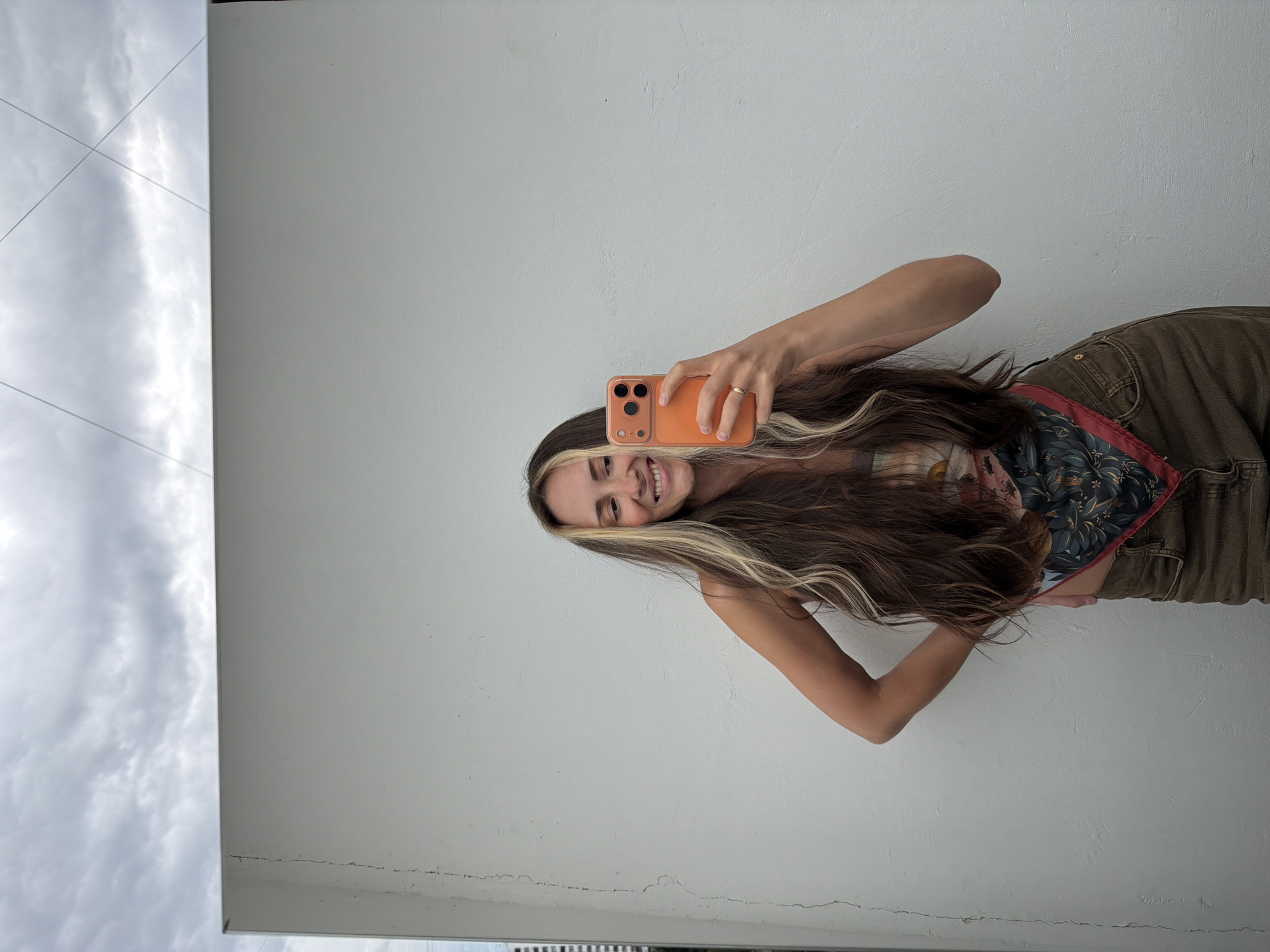 A young woman with long, wavy brown hair with blonde highlights is taking a selfie in a bright, airy setting. She is wearing a patterned tank top and khaki pants. Her right arm is extended, holding an orange smartphone, and she is smiling warmly at the camera. A ring is visible on her right hand. The background is a plain, textured white wall, with a portion of a cloudy sky visible on the left side of the frame, suggesting an overcast day. The image is taken vertically, and the orientation of the woman indicates she may be standing or leaning. The setting appears to be outdoors or near a window, given the natural light and the sky reflection. No text is visible in the image.