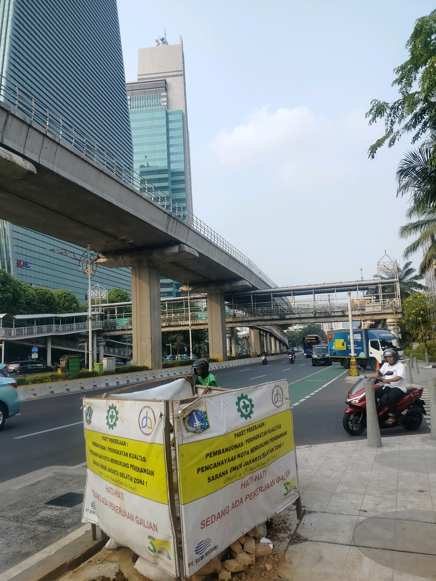 The image depicts a street scene in South Jakarta, Indonesia, likely on a clear day. In the foreground, a temporary construction barrier with yellow and white signage is positioned on the sidewalk. The signage indicates ongoing work, with text such as "PAKET PEKERJAAN: PEMBANGUNAN / PENINGKATAN KUALITAS" (Work Package: Development / Quality Improvement) and "HATI-HATI SEDANG ADA PEKERJAAN GALIAN" (Caution: Excavation Work in Progress).
Behind the barrier, a multi-lane road is visible with traffic. Cars and motorcycles are present, with one motorcycle rider in the immediate foreground wearing a white shirt and helmet. To the left of the road, a pedestrian overpass or elevated walkway system curves into the distance, supported by concrete pillars.
Dominating the background are modern skyscrapers, characteristic of a major urban center. One building on the left is a tall, glass-facaded structure with horizontal lines. Another building behind it is lighter in color and appears to have a more stepped architectural design. Palm trees and other lush greenery are interspersed with the urban landscape, particularly on the right side of the image. The sky is a pale blue with scattered clouds. The overall impression is of a busy urban environment with ongoing infrastructure development.