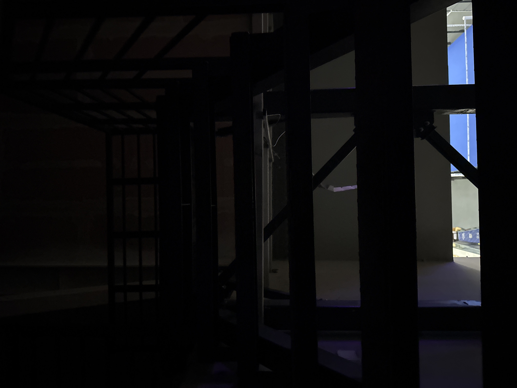 A dimly lit, silhouetted view of an industrial-style staircase with metal railings and beams. Below the staircase, a bright blue surface, possibly a tarp or pool cover, is visible, with some white scaffolding structures. The scene is dark and appears to be an interior space with some ambient light creating shadows and highlighting certain areas. The location in Yekaterinburg, Russia, is not determinable from the visual information provided in the image.
