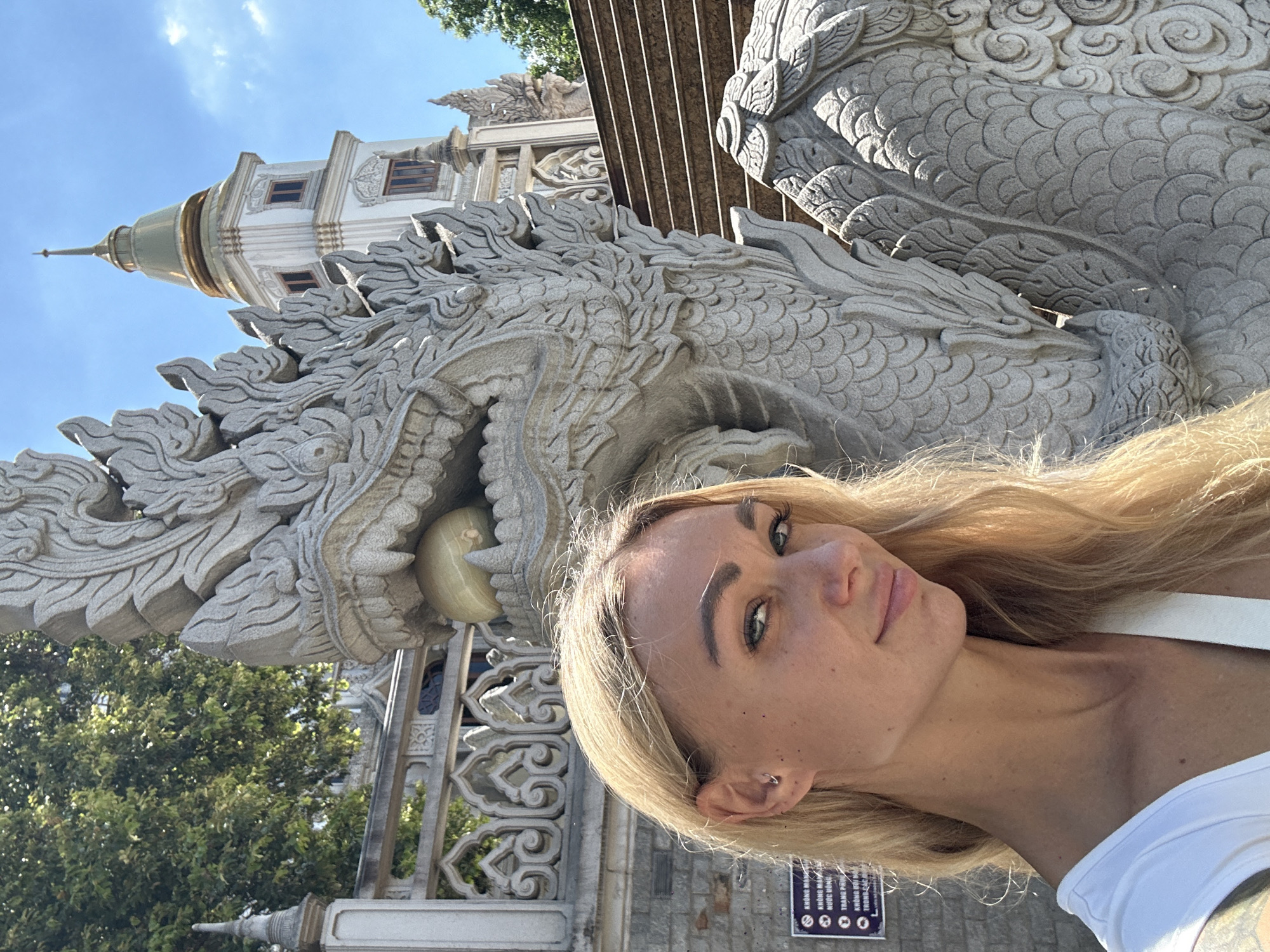 A woman with blond hair stands in front of a large stone dragon statue, with a temple structure visible in the background. The location is Dĩ An, Vietnam, as indicated by the Vietnamese language sign on the lower right. The woman is wearing a white top and has a piercing in her ear. The temple is intricately designed with a tiered roof structure. Trees are visible, suggesting a partially outdoor setting. The woman is facing the camera while holding the camera herself.