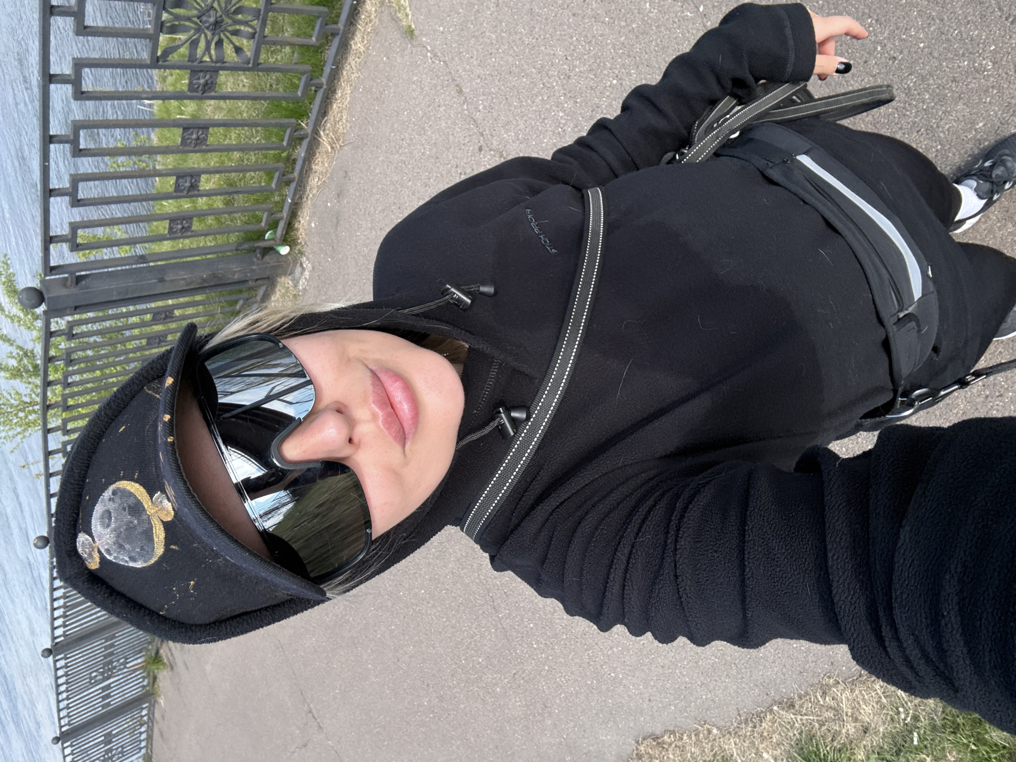A woman is taking a selfie outdoors. She wears a black hooded jacket, a fanny pack, sunglasses, and a hat decorated with moon and stars. She stands on a paved path next to a metal fence overlooking a body of water. Given the location of Krasnoyarsk, Russia, the water body is likely the Yenisey River. Grass is visible between the path and the fence. The woman's nails are painted black. She is looking directly at the camera. Her clothing is entirely black, including her shoes. The path is concrete.