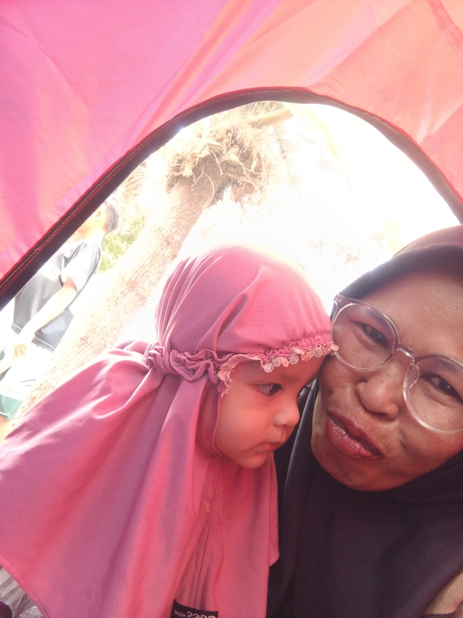 The image captures an indoor selfie taken inside a tent, likely in Perbawati, Indonesia. The foreground is dominated by two individuals. On the right, a woman wearing a dark headscarf and clear-rimmed glasses is looking towards the camera, with her face partially obscured. Her lips are painted with lipstick. To her left, a young child wearing a pink hijab with ruffled trim is positioned closer to the camera. The child's face is illuminated by warm light and their gaze is directed downwards. Behind the subjects, the bright interior of the tent is visible, along with an opening revealing a sun-drenched outdoor scene. Through the opening, a large tree trunk and foliage can be discerned. A blurred figure wearing a gray t-shirt and green shorts is partially visible in the background. The overall scene suggests a moment of familial bonding or a casual outing in an outdoor setting.