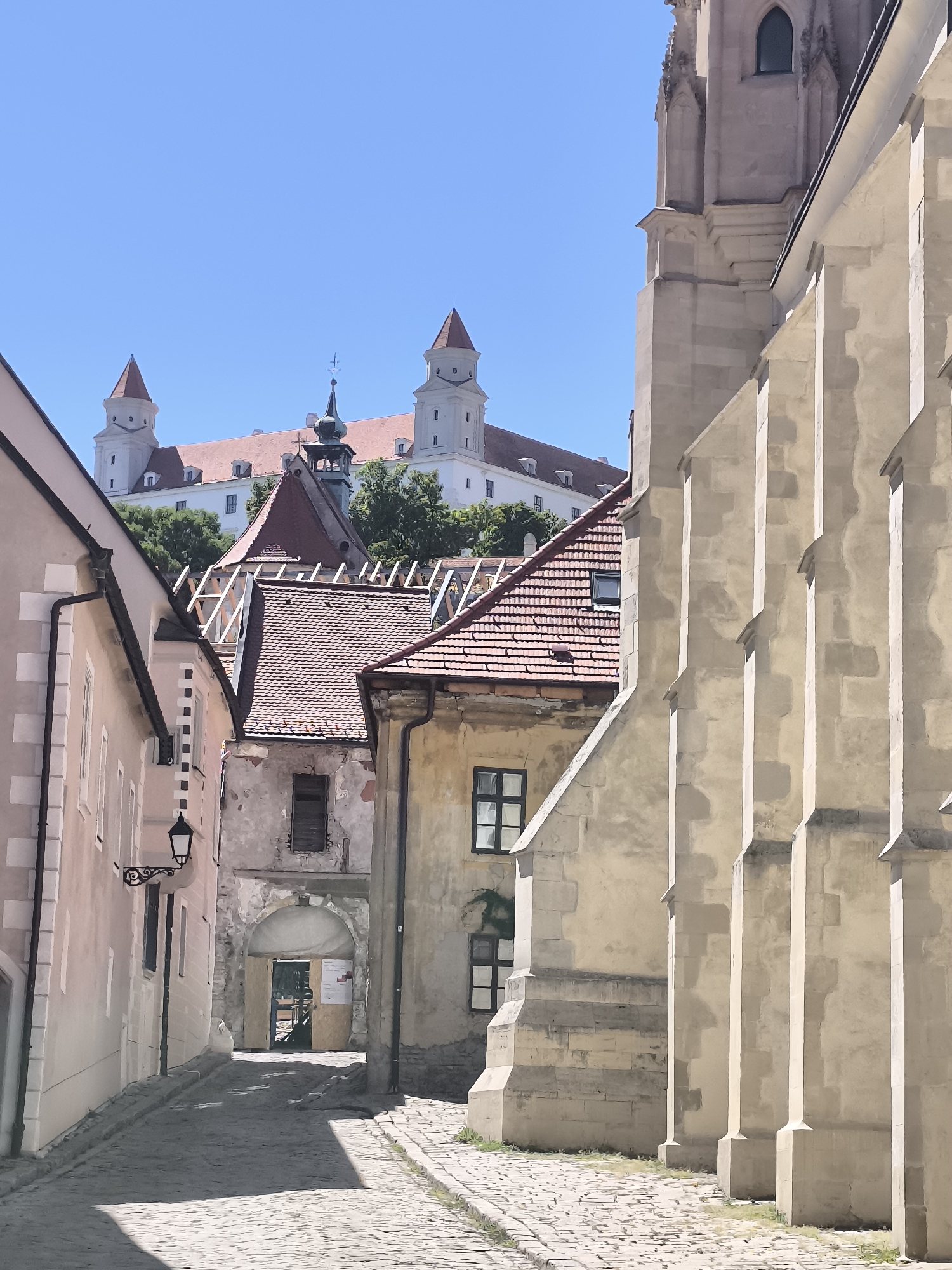 The image shows a narrow, cobblestone street in Bratislava, Slovakia, leading towards Bratislava Castle, which is visible in the background atop a hill. The castle is a large, white structure with red-tiled roofs and several towers, including two prominent ones with conical roofs. The street is lined with old buildings of varying architectural styles and colors. On the left, a pastel-pink building features a black wrought-iron lamppost. To the right of the street, a large, light-colored stone building with buttresses dominates the foreground, likely part of a church or cathedral. Several red-tiled roofs of smaller buildings are visible between the larger structures. The sky is clear and blue. There are no people or animals visible in the scene. The overall impression is one of historical architecture and a sunny day in an old European city.