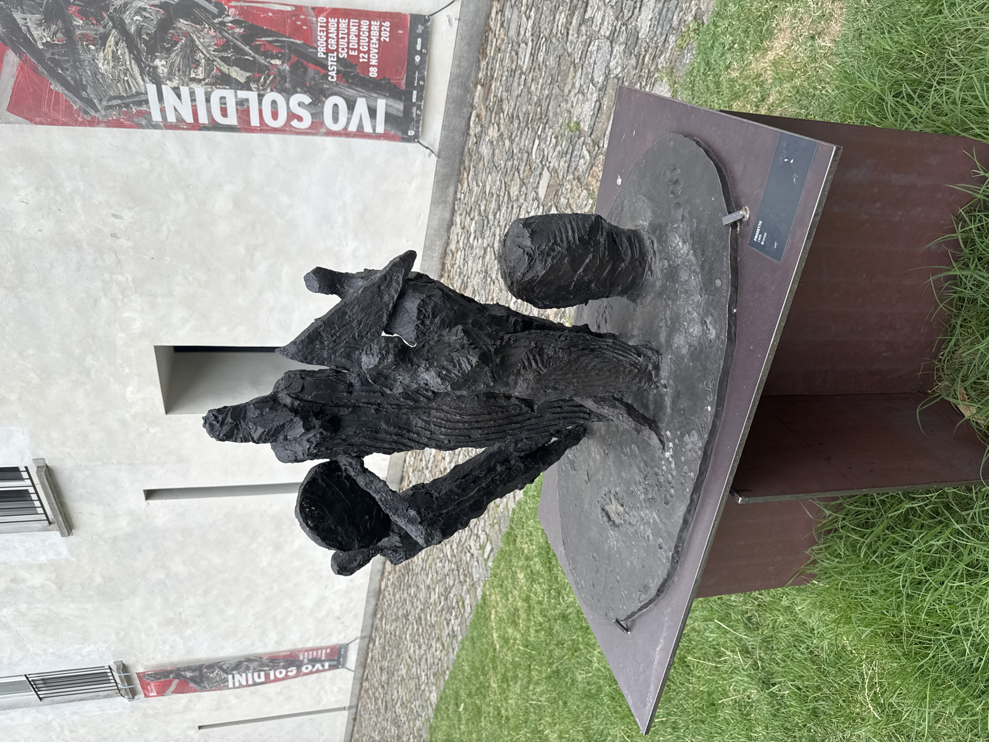 The image shows a dark bronze sculpture, characterized by abstract human-like figures and geometric shapes, resting on a dark metal pedestal. The sculpture appears to be placed outdoors, with a cobblestone ground and green grass surrounding the display. In the background, a light-colored building with windows and a balcony is visible, adorned with two large red banners featuring black text and imagery. The banners are indicative of an art exhibition or event. The overall scene suggests an outdoor art installation in a public or historical setting. Based on the text on the banners, which includes "IVO SOLDINI" and "PROGETTO CASTEL GRANDE SCULTURE E DIPINTI 12 GIUGNO - 08 NOVEMBRE 2026", the location is Castel Grande in Bellinzona, Switzerland.
