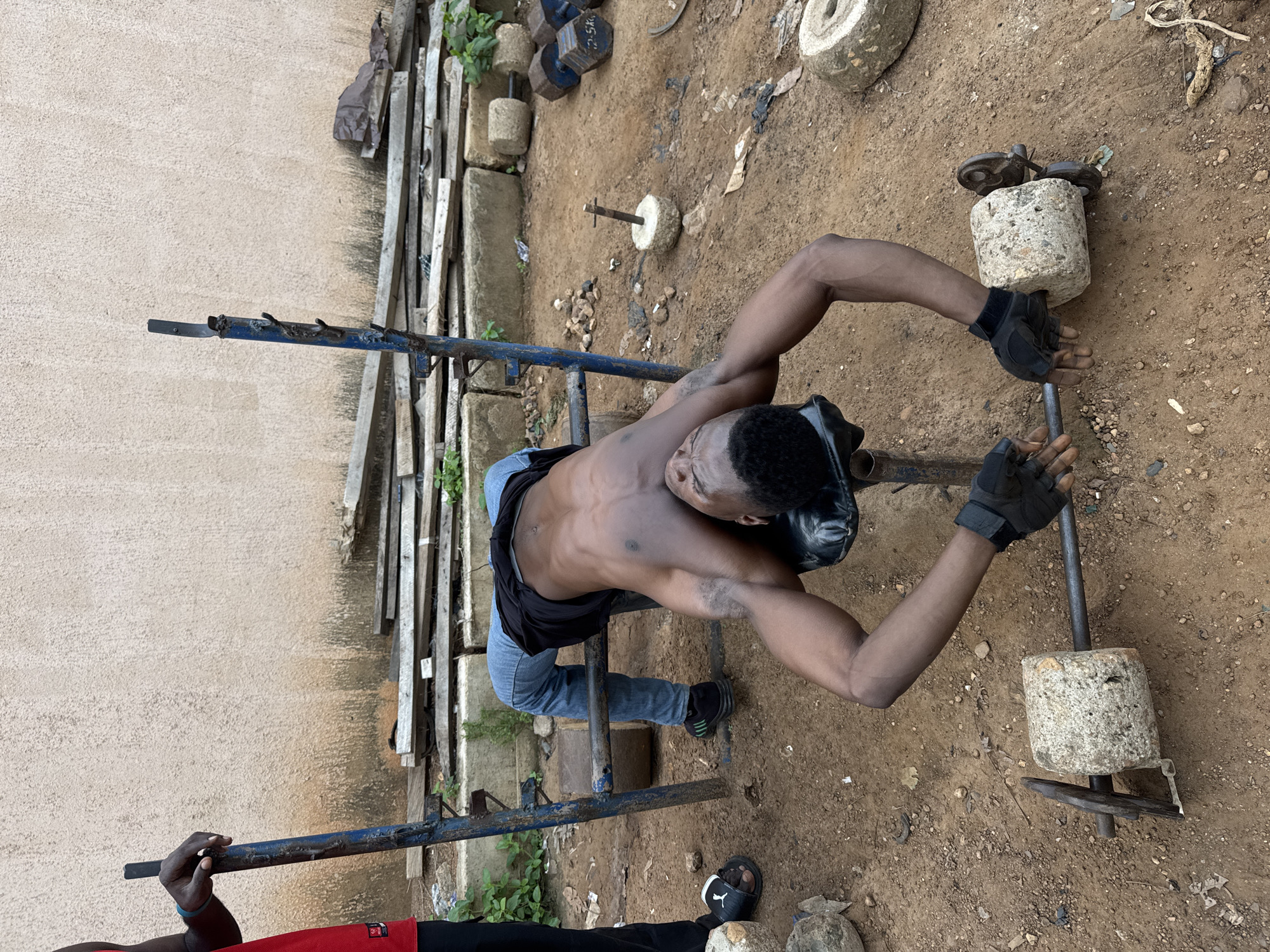 In Akwanga, Nigeria, a young Black man is engaged in a strength training exercise outdoors. He is lying supine on a makeshift weight bench, which appears to be constructed from aged blue metal poles and a black upholstered surface. The bench is situated on uneven, dirt ground, littered with small stones and debris.
The man is wearing blue jeans and a black t-shirt, with black gloves on his hands. He is holding a barbell with two weights made of concrete, each appearing roughly square with circular indentations. One concrete weight is visible on the left side of the barbell and another on the right. He is in the process of lifting the barbell, with his arms extended and his body positioned to perform a chest press or similar exercise. His musculature is clearly defined, indicating a dedicated fitness regimen.
In the background, there is a rough-textured, light-colored wall. Piles of stacked wooden planks and various stones, some appearing to be repurposed weights or construction materials, are scattered around the area. To the left of the frame, another Black individual, partially visible, stands near the weight bench, their arm resting on one of the blue metal supports. The overall environment suggests a rustic, improvised gym setting.