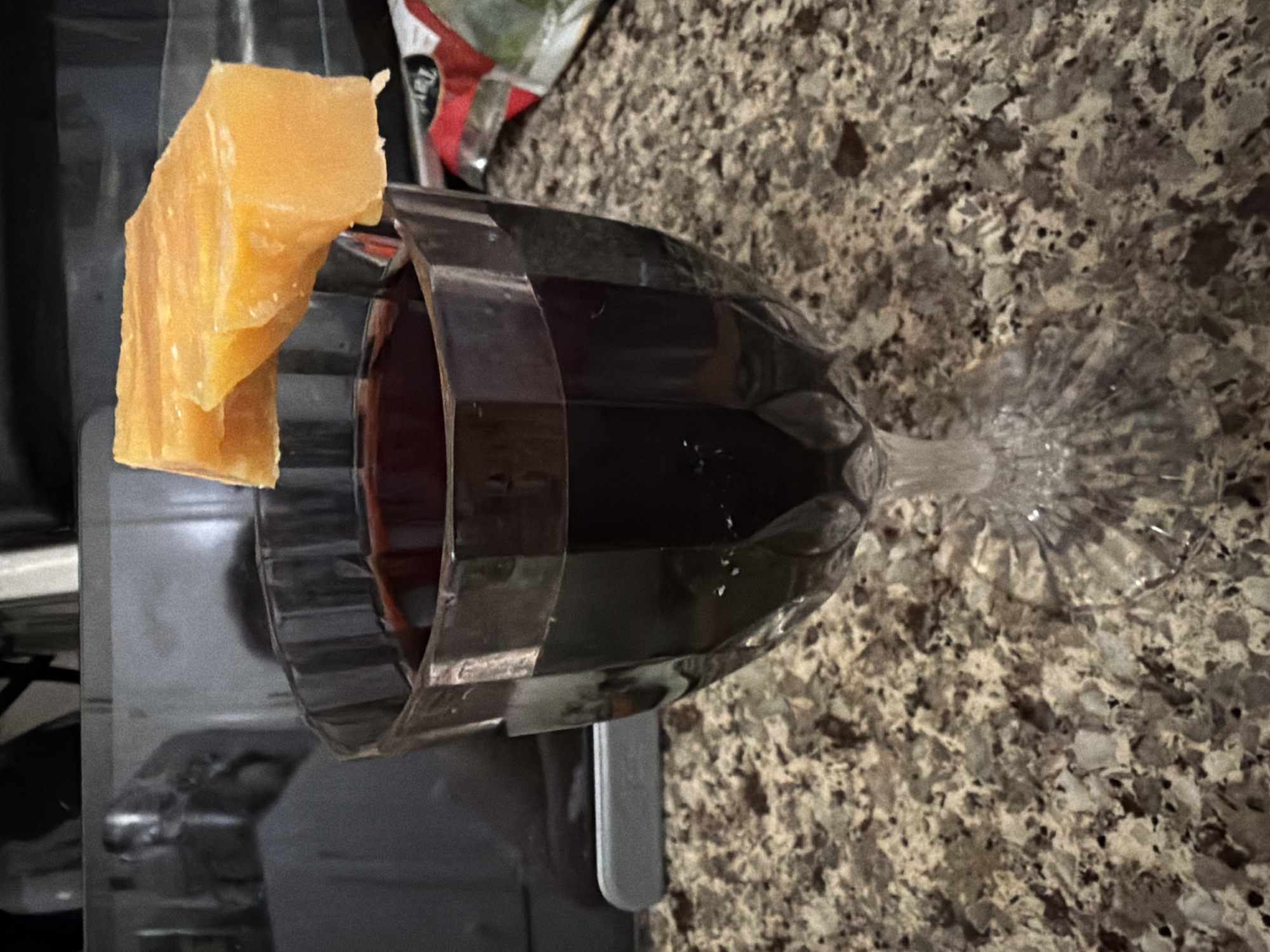 A close-up, slightly overhead shot focuses on a faceted glass goblet filled with dark red wine. The goblet is placed on a speckled granite countertop. Resting precariously on the rim of the glass is a rectangular piece of yellow cheese, appearing firm and slightly translucent. In the blurred background, a dark appliance and some packaging with red and white accents are visible. The scene is indoors, likely in a domestic kitchen.