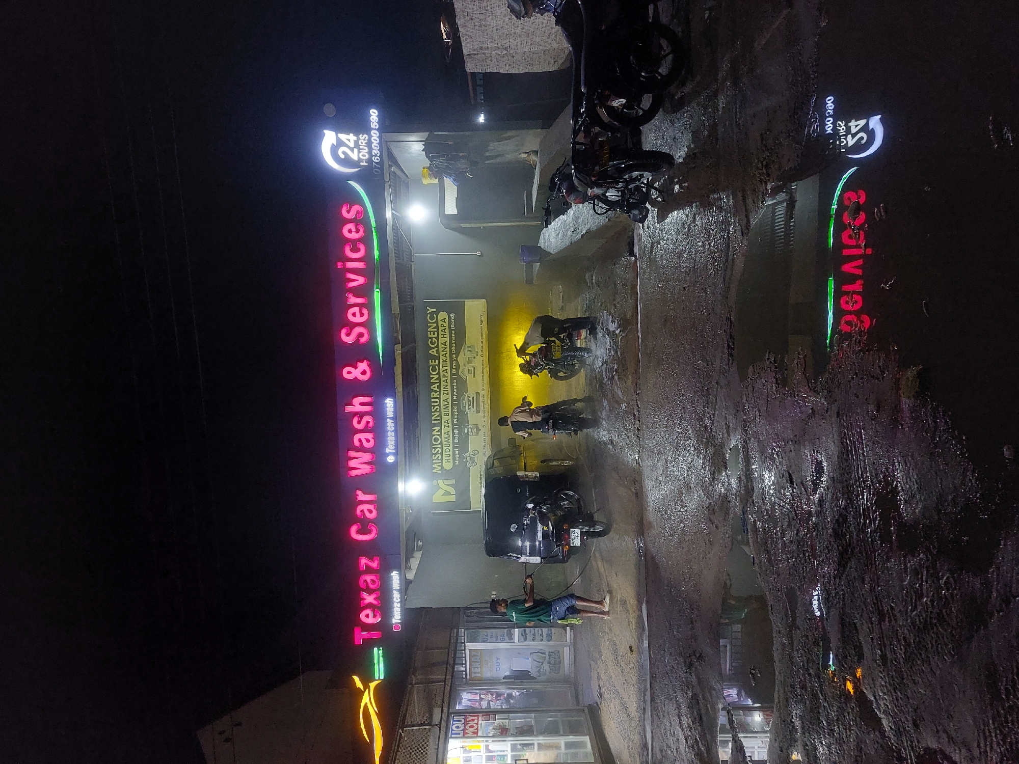 The image shows a car wash business at night in Dar es-Salaam, Tanzania, called "Texaz Car Wash & Services." The name is displayed in pink neon, next to a yellow car logo. There's a "24 hours" sign above the car wash name and an insurance company billboard. A man is washing a motor tricycle, while other motorbikes are nearby. The road looks wet. The business is lit by fluorescent lights. The car wash also sells motor oil. There is another neon sign for a similar car wash across the street.