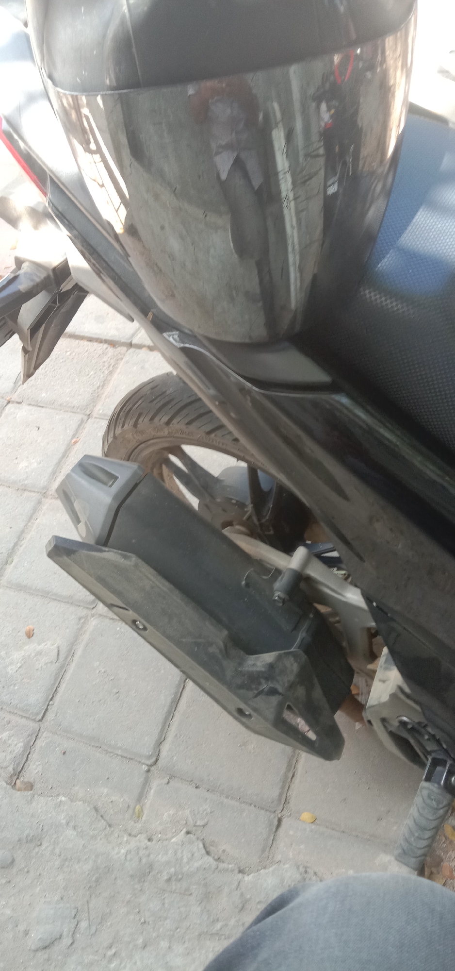 The image displays a close-up, slightly angled view of the rear portion of a black motorcycle parked on a paved surface. The motorcycle's black seat is visible, as is a portion of its black exhaust system and rear tire. The surrounding environment appears to be an outdoor urban setting, likely a street or sidewalk in Kendari, Indonesia, suggested by the textured concrete paving stones. The lighting indicates daytime. No individuals are directly visible in the frame, but a person's leg, clad in grey fabric, occupies the bottom portion of the image, suggesting a person is either standing or sitting near the motorcycle. The motorcycle exhibits signs of use, with some dust and minor scuffs visible on its surfaces.