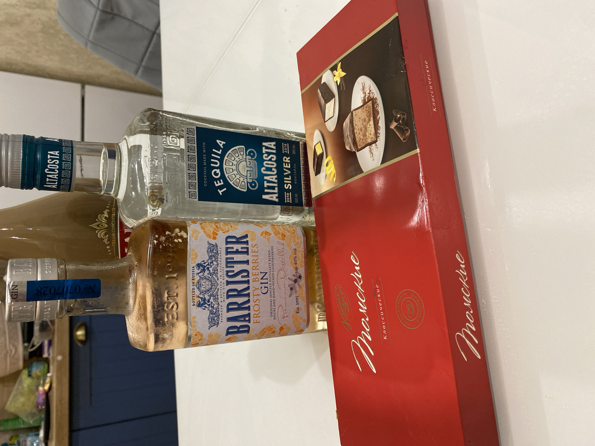 This image captures an indoor scene in the city of Tyumen, Russia, featuring alcoholic beverages and a box of confectionery items arranged on a white table.
In the foreground, a red rectangular box is positioned, displaying the brand name "Красная Заря" (Krasnaya Zarya) and the product name "Томские Классические" (Tomskie Klassicheskie), translated as "Tomsk Classic." The box features an image of three chocolate-covered desserts, possibly a type of soufflé or cake bar, garnished with cocoa powder, lemon zest, vanilla, and cinnamon sticks. The phrase "РУЧНАЯ РАБОТА С ЛЮБОВЬЮ" (Handmade with love) is also visible.
Behind the box, two clear glass bottles containing alcoholic beverages are present. The bottle on the left is Barrister Frosty Berries Gin, labeled "BOTTLED IN RUSSIA" and "FROSTY BERRIES GIN," indicating it's an "ORIGINAL JUNIPER DISTILLATE BLEND, SPICES AND HAND-PICKED CLOUDBERRIES." It states "Premium Distilled," "Est. 1995," and "40% vol." A blue excise stamp with "№ 0707028" is affixed to its neck.
The bottle on the right is AltaCosta Tequila Silver. Its label reads "COCKTAIL MADE WITH TEQUILA," "ALTACOSTA SILVER," "ESTD 2018," and "38% Alc," with a volume of "500 ML" and the word "КОКТЕЙЛЬ" (Cocktail).
In the background, a kitchen or dining area is visible. To the left, dark blue kitchen cabinets with a gold-colored handle are seen, topped by a light wood-colored countertop on which various blurred kitchen items and containers are placed. A beige/brown bottle, partially visible between the two main bottles, has "CREAM CONCENTRATE" printed on its label. To the right, parts of a white refrigerator and a grey upholstered chair are discernible against a light wall. No individuals are present in the frame.