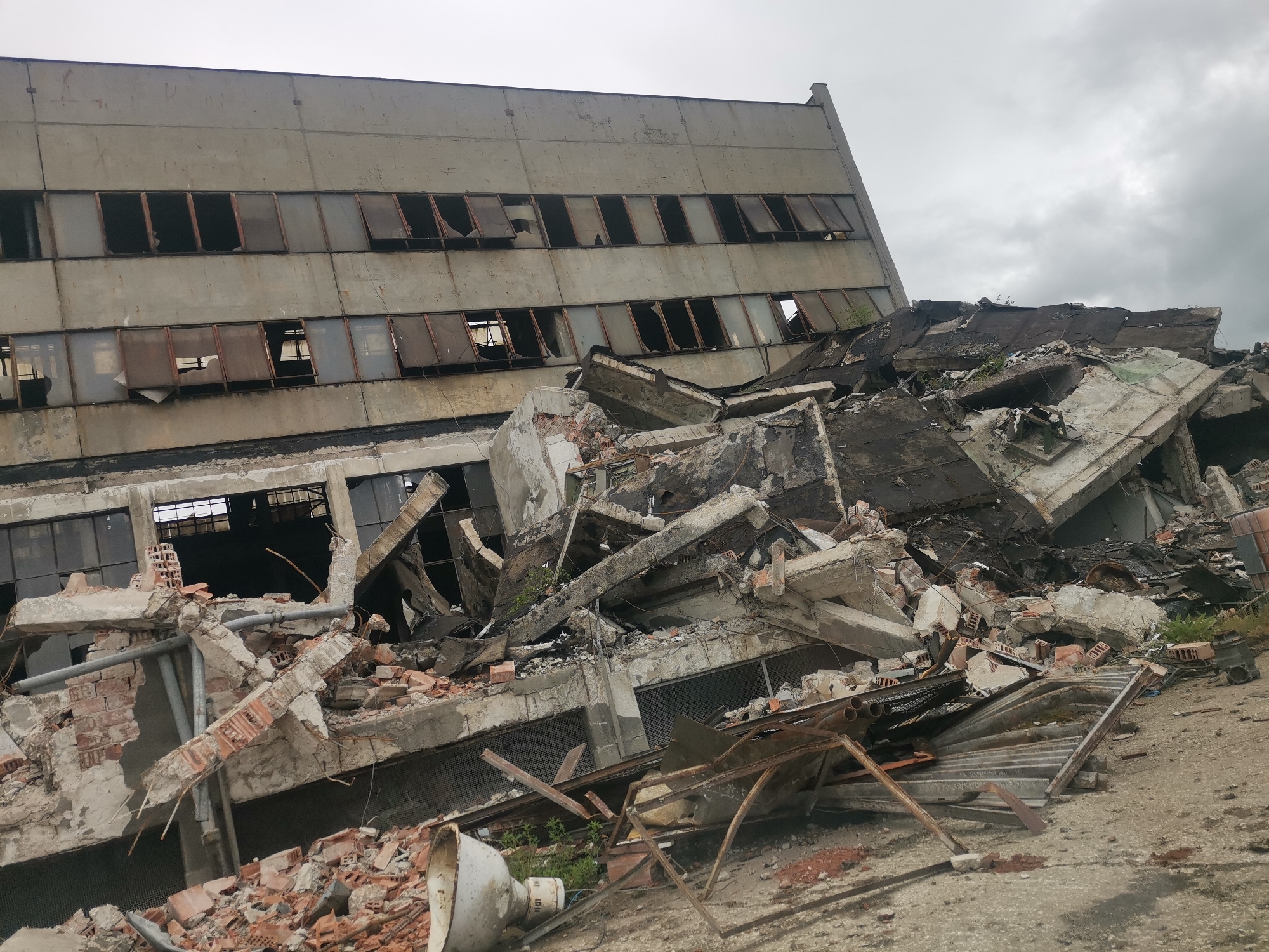 The image shows a partially collapsed, multi-story concrete building in Zhivkovo, Bulgaria. The building has multiple rows of rectangular window openings, many with missing or broken glass. A large pile of rubble consisting of broken concrete, bricks, metal, and other debris is visible in front and on top of the building's remaining structure. The debris obscures much of the lower portion of the building. The sky is overcast. A white horn speaker lies amidst the rubble in the lower left corner of the image.