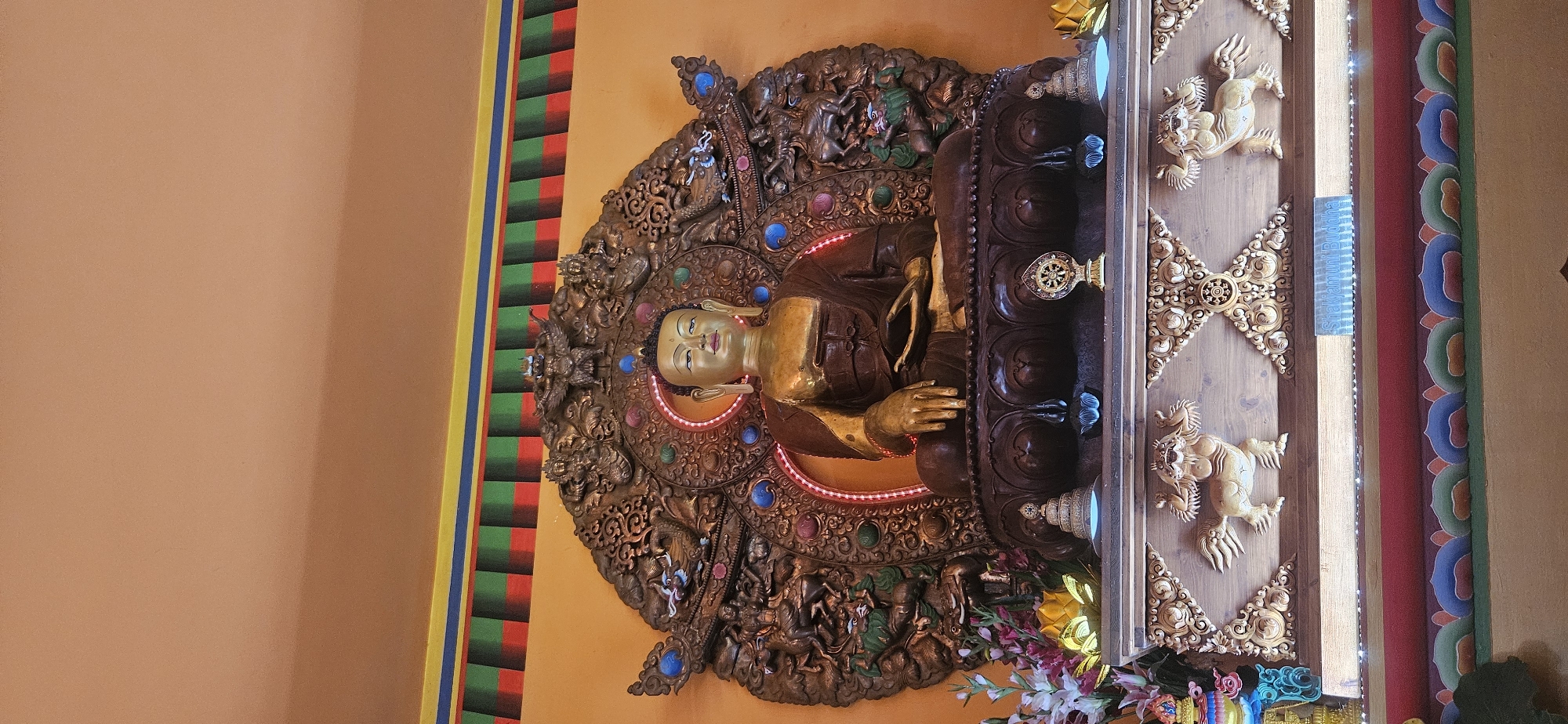 The image displays a large, golden statue of Buddha seated in meditation on a dark, ornate base. The statue is positioned centrally, set against a warm orange wall. Behind the Buddha, a large, intricately carved golden aureole frames the figure. This aureole is adorned with numerous detailed figures, including what appear to be dragons and mythical creatures, interspersed with colorful, jewel-like accents. Above the aureole, a decorative frieze runs along the wall, featuring bands of yellow, green, red, and blue.
The Buddha statue is gold-colored with a darker brown robe draped over its shoulders. Its hands are resting in its lap, in a meditative pose. The facial features are serene and detailed. The base supporting the Buddha is dark brown and elaborately carved, featuring lotus petal motifs.
Below the main statue, a wooden altar is visible. This altar is decorated with carved figures of mythical beasts and ornate patterns. Various smaller objects are placed on the altar, including flowers (some appearing artificial), small stupa-like structures, and a Dharma wheel symbol. A small plaque with the text "Shakyamuni Buddha" is affixed to the front of the altar. The overall environment suggests a temple or spiritual site. The location of this statue is the Béke Sztúpa (Peace Stupa) in Zalaszántó, Hungary.