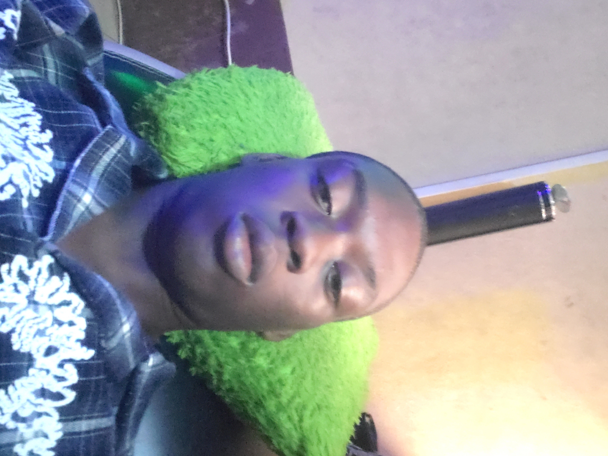 A man is lying down against a fluffy green pillow. He is wearing a blue and white plaid shirt with white floral embroidery. A dark, cylindrical object with a silver tip is visible in the background. The location is Unknown, Unknown.