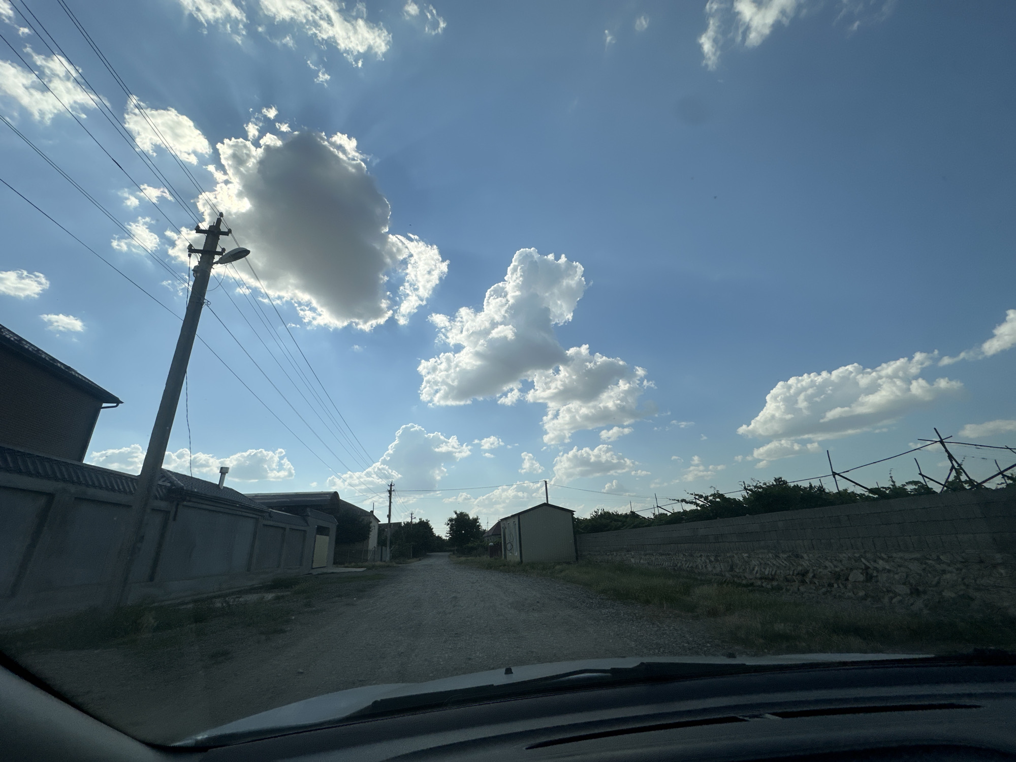 The image depicts a view from inside a vehicle looking out at a rural road. The sky is blue with scattered white clouds, some of which are illuminated by the sun, creating rays of light. A power line pole with attached wires and a street lamp stands to the left of the road. Along the left side of the road, there is a grey wall and a building. The road itself is unpaved and appears to be gravel. Further down the road, there are more buildings and trees. On the right side of the road, there is a stone wall and vegetation. The overall scene suggests a quiet, possibly remote, location on a sunny day. The location is identified as Subutli-Miatli, Russia.