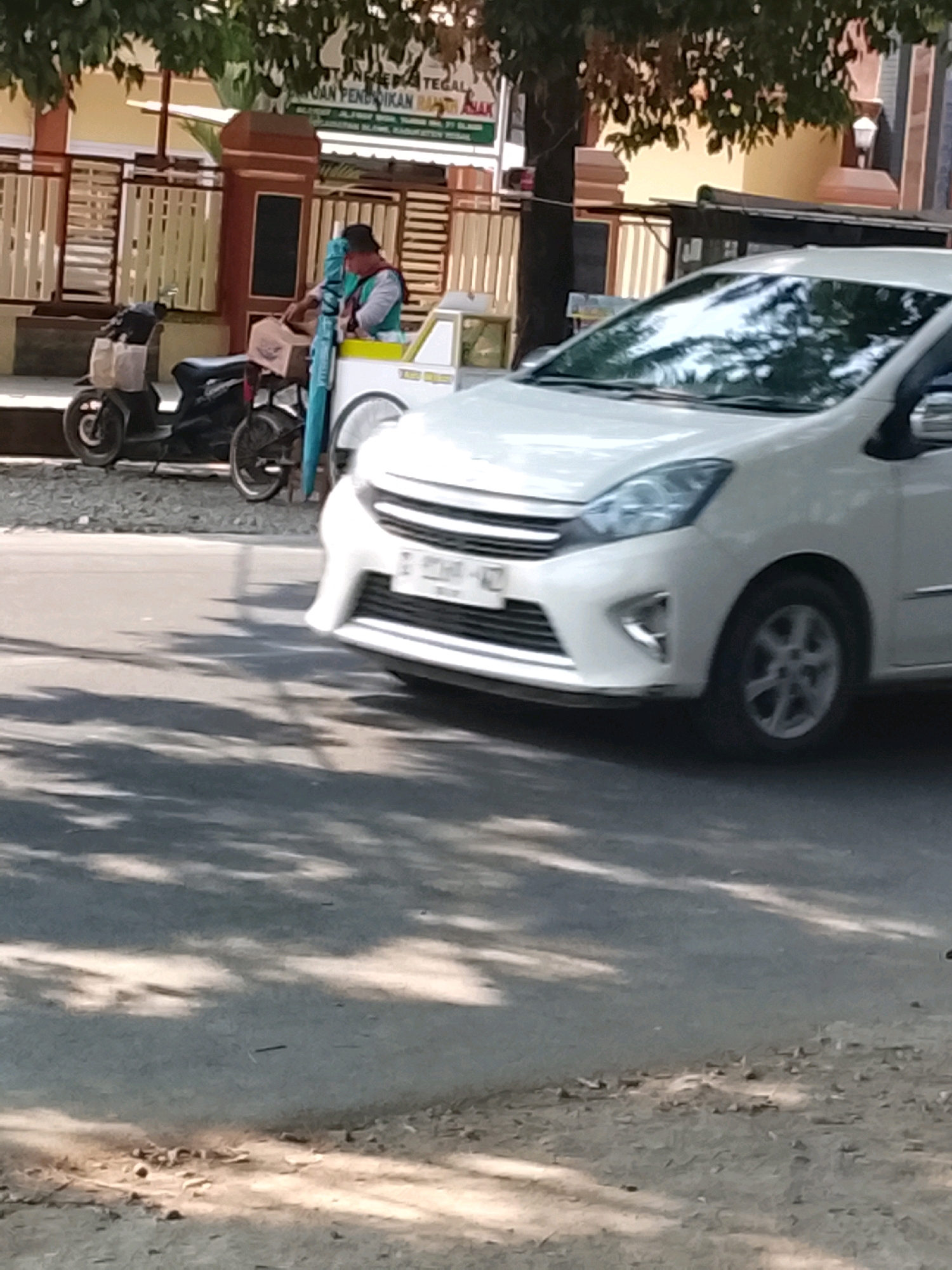 In the city of Slawi, Indonesia, a white Toyota Avanza car is parked on the side of the road, angled towards the camera. The car's front right wheel is visible, and its license plate reads "G 8214 WZ". To the left of the car, a person wearing a green shirt and a dark head covering is standing next to a white cart, possibly a food or beverage vendor. Behind the vendor, a black motorcycle is parked. The scene is illuminated by sunlight, casting shadows on the road and ground. In the background, there is a building with a fence and some trees.