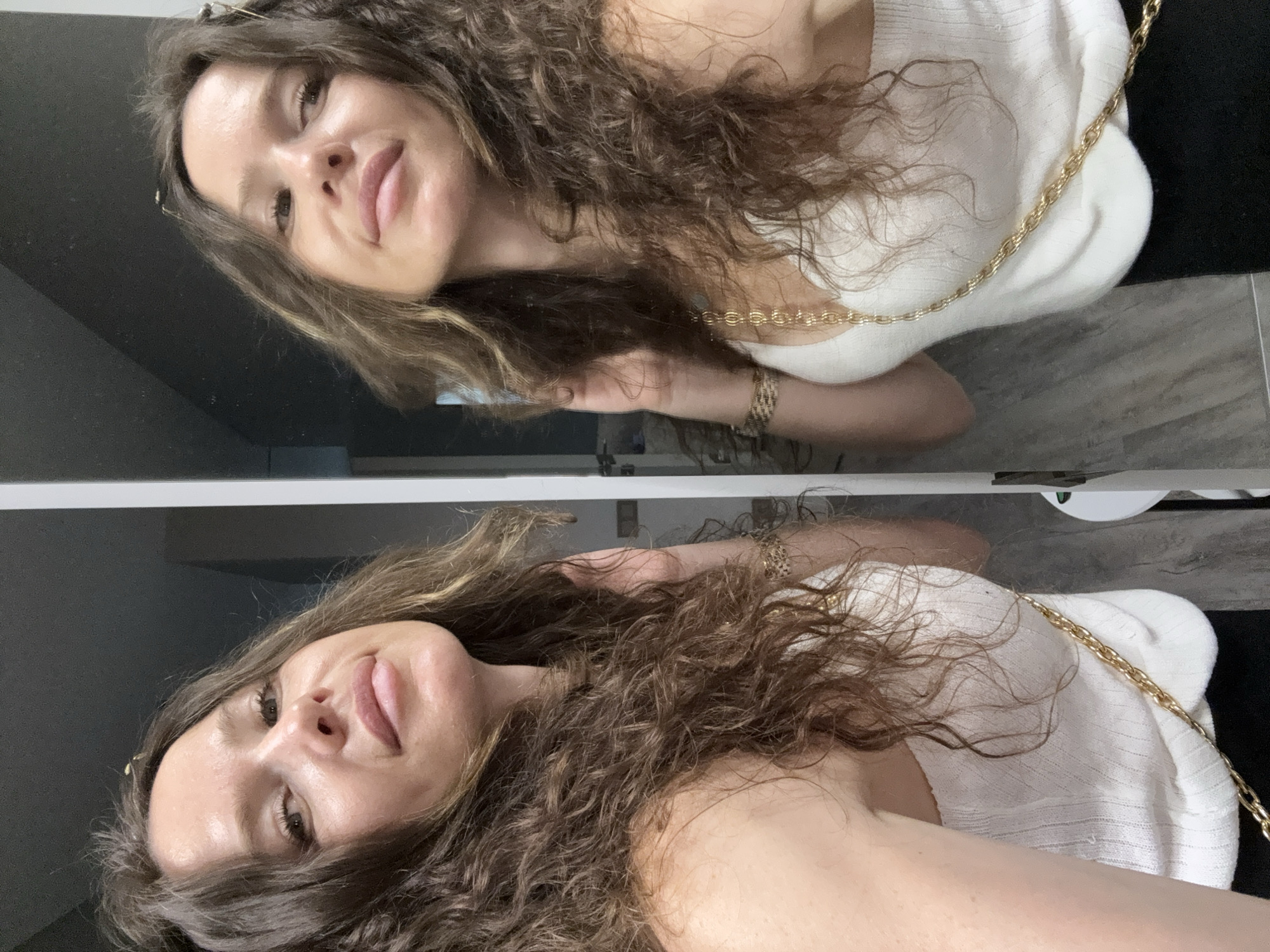 A woman with long, wavy brown hair and fair skin is looking into a mirror. She is wearing a white ribbed tank top with a gold chain strap and a black skirt. Her left hand is resting on her hair near her face, and her right hand is on her hip, with a gold bracelet visible on her wrist. The reflection shows her from a slightly different angle. The background is a dark, modern interior, possibly a bathroom or dressing area, with a tiled floor.