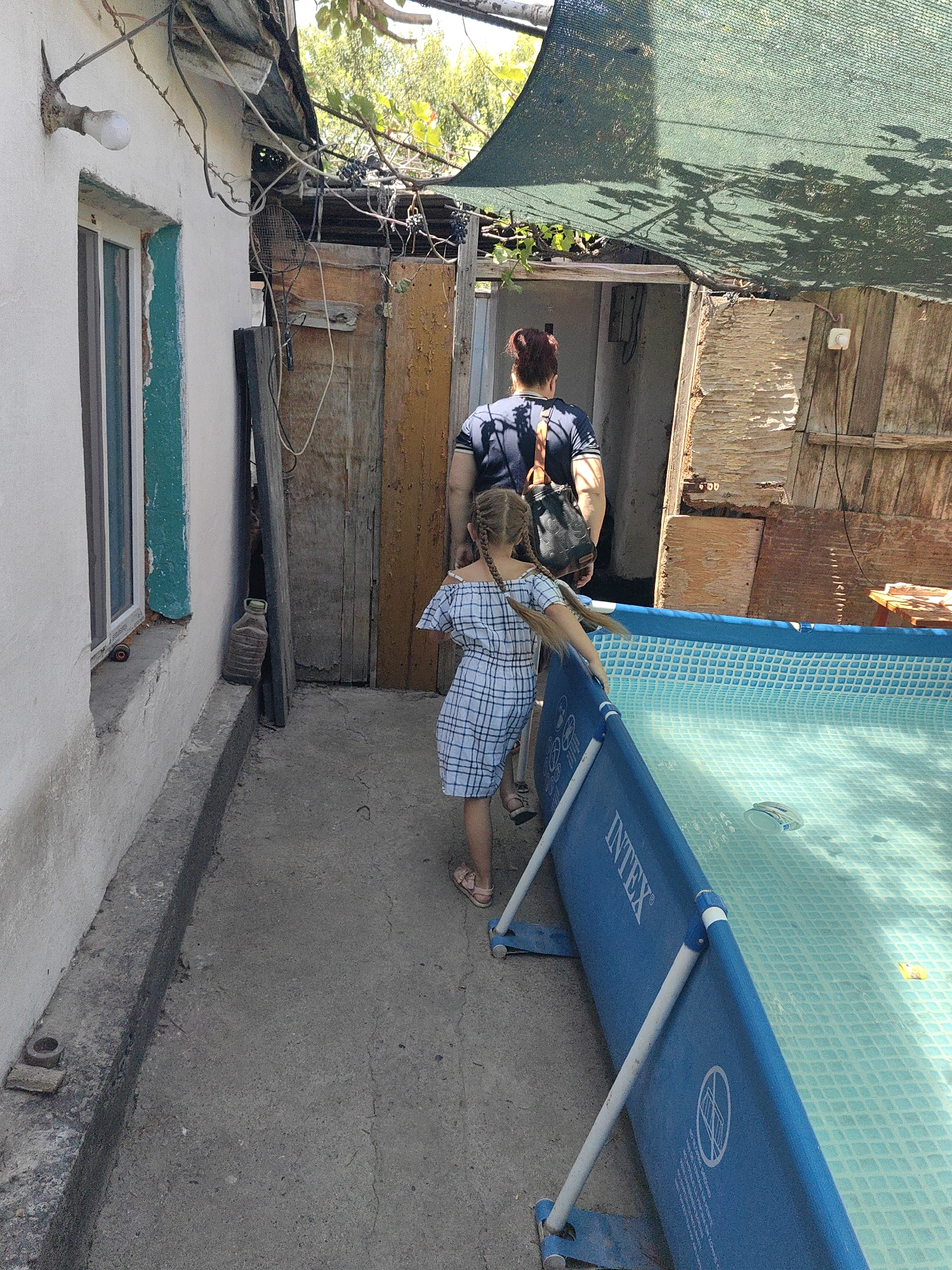 A woman with auburn hair tied back, wearing a dark t-shirt and a crossbody bag, walks away from the camera towards an open doorway. Beside her, a young girl with braided blonde hair, wearing a blue and white checkered off-the-shoulder dress, walks towards a blue, inflatable swimming pool. The girl's right hand is touching the side of the pool. The swimming pool, labeled "INTEX," is filled with clear water. The environment appears to be a residential backyard with a concrete pathway. To the left of the pathway is a white-painted wall with a window and an outdoor light fixture. To the right, there is a wooden structure and a green mesh canopy overhead, with tree branches and leaves visible. The scene is set in Shymkent, Kazakhstan.