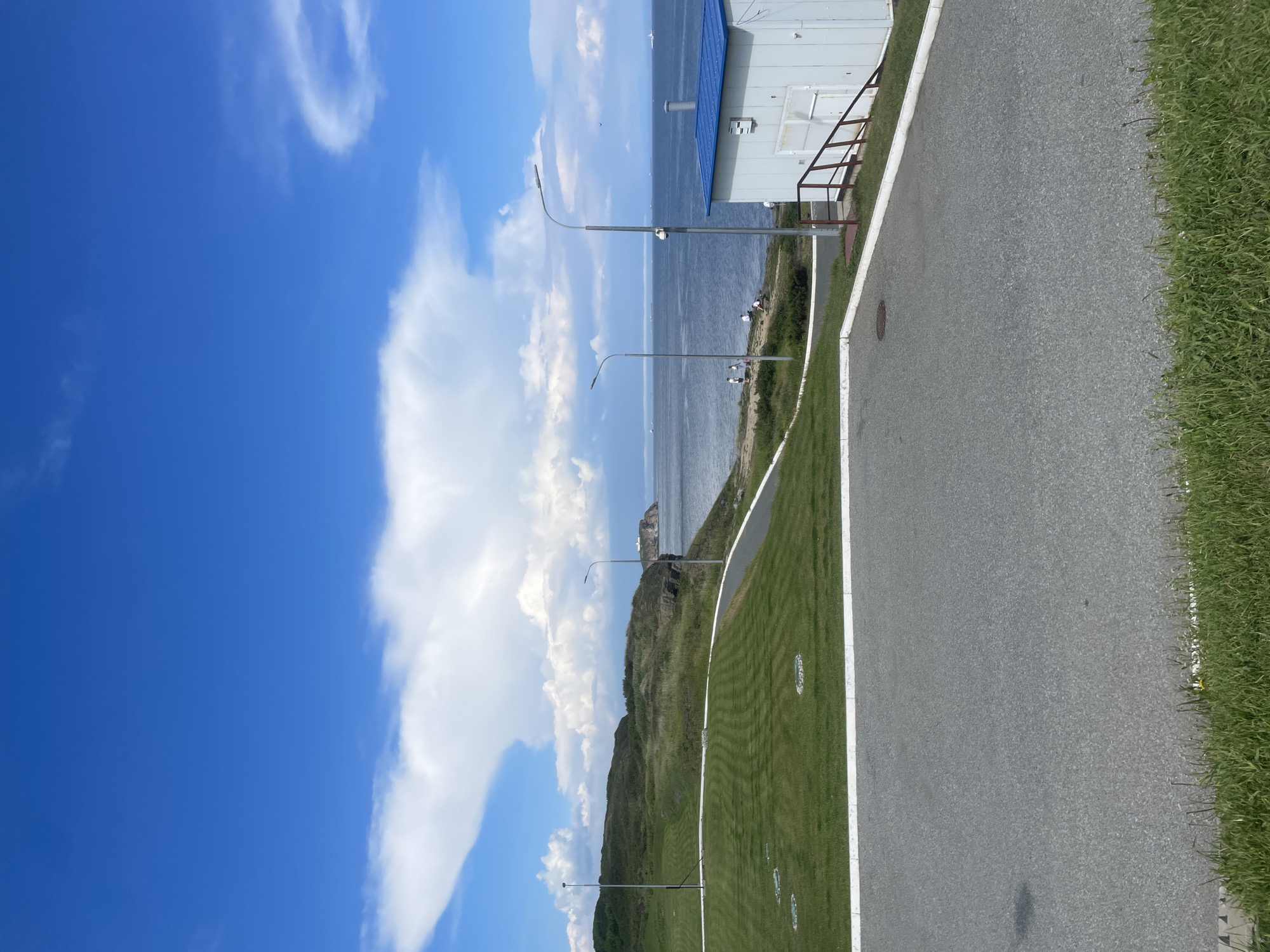 The image captures a scenic view of a coastal area on Russky Island, Russia. The foreground features a paved area and a well-maintained green lawn with distinct striping, suggesting a park or recreational space. A white building with a blue roof and a brown railing leading to its entrance is situated on the right side of the frame. Beyond the lawn, a winding paved path leads towards the coastline, where several individuals are seen gathered near the water. The sea stretches out towards the horizon, dotted with numerous ships. A rugged, green-covered hill rises from the coast, with a small lighthouse-like structure visible on a distant island. The sky is a vibrant blue, adorned with large, fluffy white clouds, creating a dramatic contrast. The overall scene conveys a sense of peacefulness and natural beauty, characteristic of a day at the seaside.