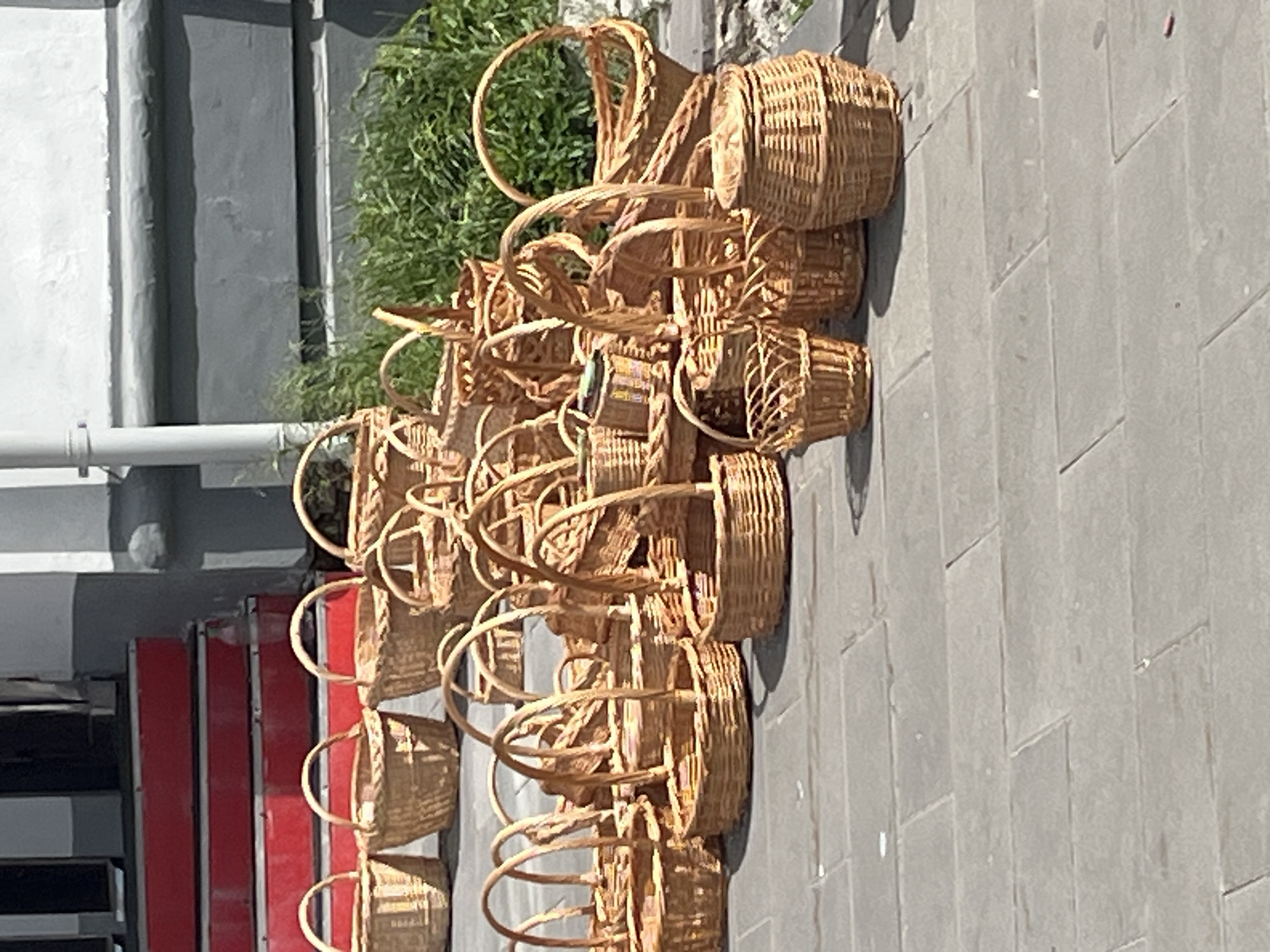 The image shows a collection of approximately 15-20 wicker baskets of varying sizes. They are stacked or arranged in a row on a light grey paved surface. In the background there is a wall, and a small area of green vegetation. A red and black metal structure is also partially visible in the lower-left corner. The location is Izhevsk, Russia.
