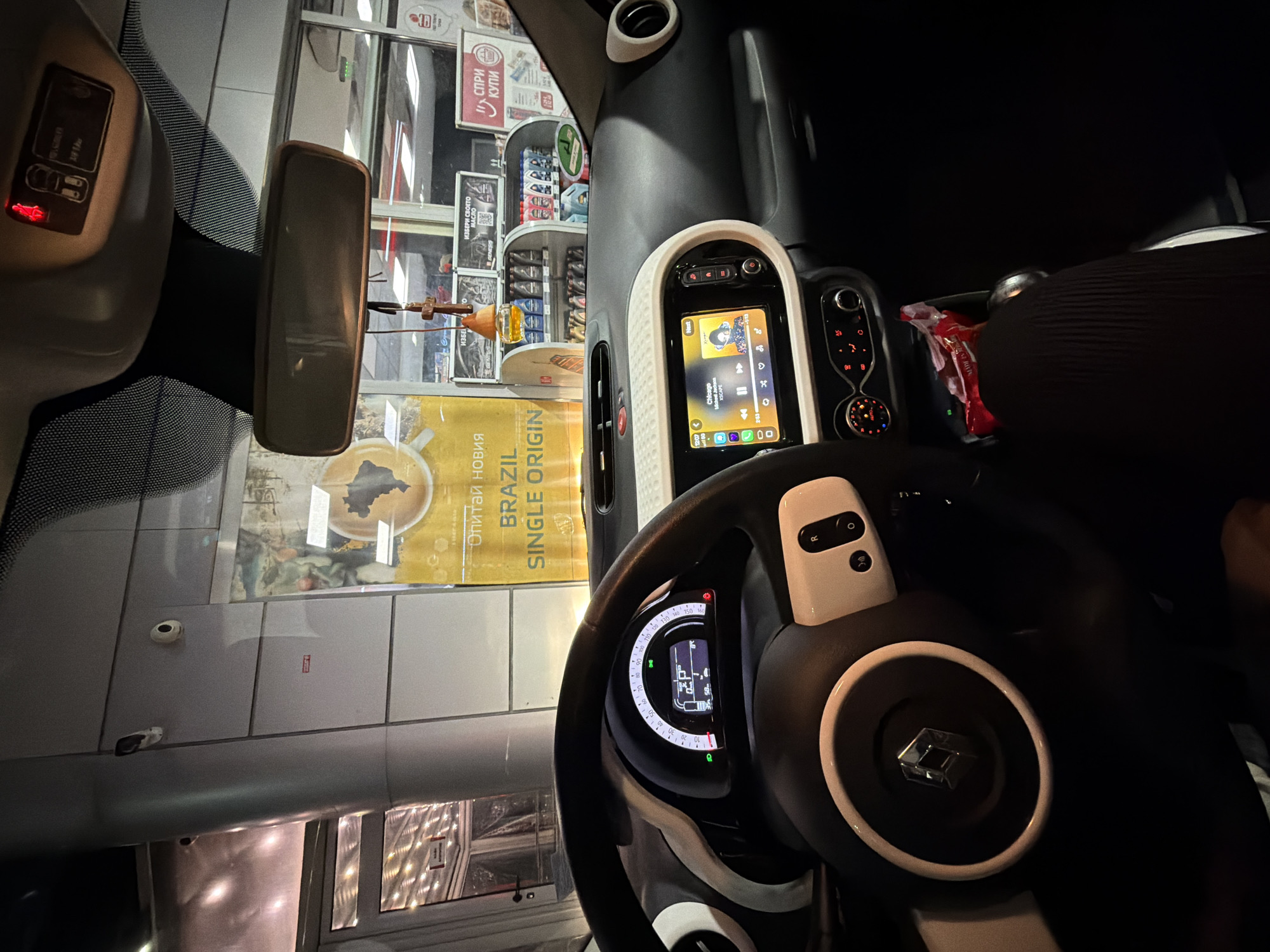 The image depicts the interior of a car, specifically the driver's seat and dashboard area. The steering wheel is prominently featured, showing the Renault logo. The car's infotainment system displays music information. Through the windshield, an advertisement for "Brazil Single Origin" coffee is visible on a storefront, along with other store displays. Based on the visible text and product selection, this is most likely a gas station. The setting seems to be Gorni Bogrov, Bulgaria.
