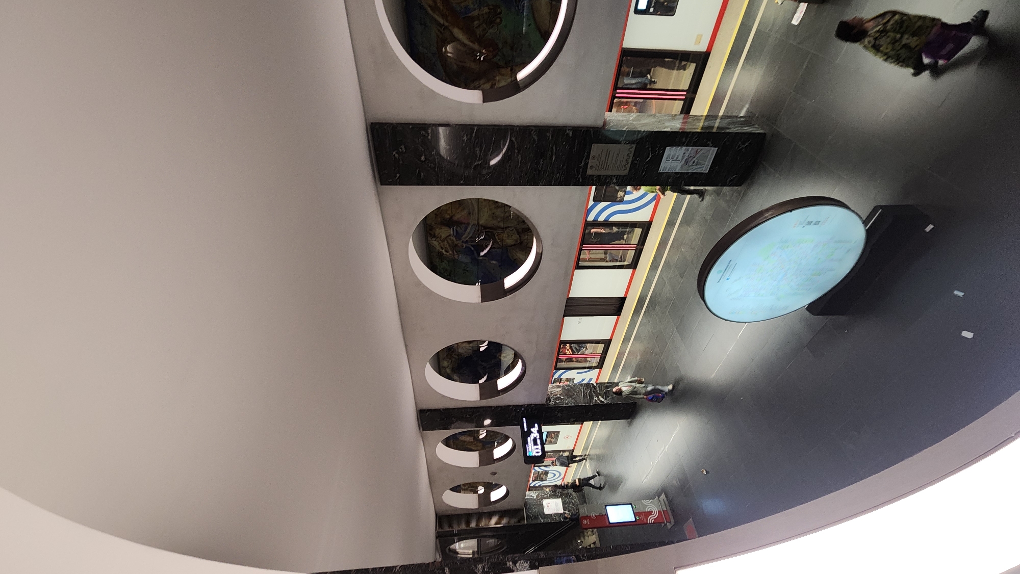 This image captures the interior of a modern, well-lit underground subway station, likely part of the Moscow Metro system. The perspective is angled, looking across the platform towards a stopped train.
The station's design is contemporary and sleek. A wide, light-colored (white or light gray) ceiling arches overhead. Along one side of the platform, a decorative wall features several large, circular cutouts, each containing intricate artistic murals or decorations, possibly mosaics. A substantial, dark-colored, rectangular pillar, appearing to be made of marble or a similar material, stands centrally on the platform.
A modern subway train is positioned along the platform tracks. Its exterior is predominantly white with distinctive blue and red wave-like patterns. Reflections of the station and passengers are visible in its windows.
The platform floor is dark gray or black, likely tiled, contributing to the clean aesthetic. Several people are present on the platform, indicating an active commute. One individual, blurred from movement, is seen walking away in the distance, wearing a patterned jacket and carrying a purple bag. Other commuters are visible further along the platform, near the train doors or standing.
A large, circular, glowing information display stands on the open platform area, showing what appears to be a metro map or informational graphic. Further down the platform, smaller digital screens are mounted, with one clearly displaying the time "07:34," suggesting the scene takes place in the early morning. Text on a plaque on the dark pillar and details on the circular map are not legible. The station environment appears functional, well-maintained, and artistically enhanced.