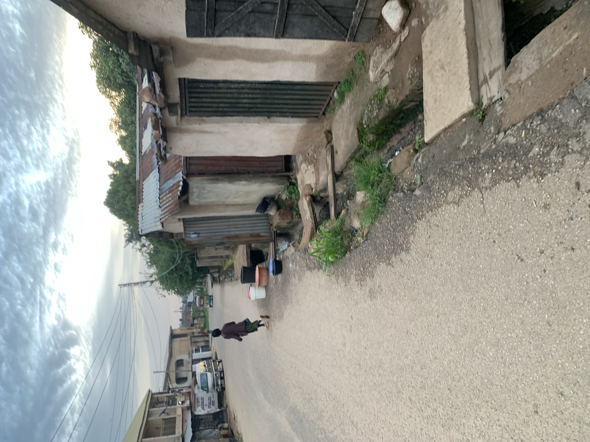 In Jos, Nigeria, a young person in a purple top and green skirt runs down a paved street. To their right, a row of small, weathered buildings with corrugated metal roofs stand adjacent to the road, some featuring metal doors. Several buckets, including a white, orange, and blue one, are placed near the buildings. In the background, a white van and a motorized tricycle are visible. The sky is overcast with light clouds.