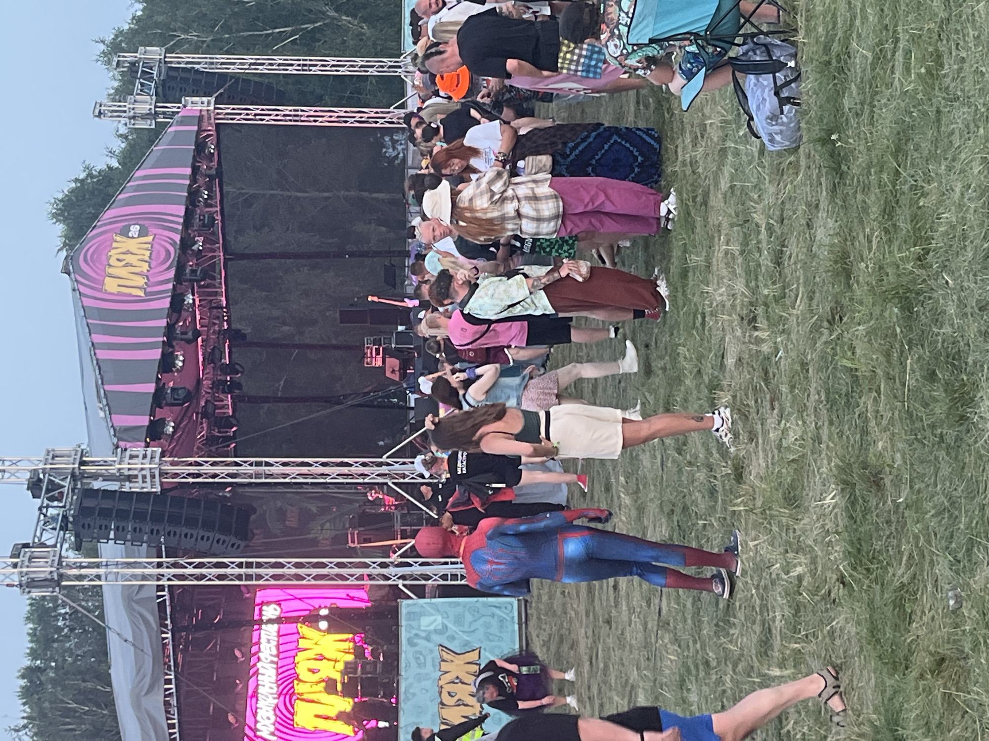 A wide shot captures a lively outdoor event, possibly a music festival, in Surakovo, Russia. The scene is dominated by a large stage structure with prominent audio equipment and bright pink and purple lighting illuminating the upper canopy. A large LED screen displays vibrant yellow text in Russian, possibly the event's name. In the foreground, a crowd of people are gathered on a grassy field. Among them, a person dressed in a full Spider-Man costume stands out, facing away from the camera. Other attendees are seen in casual attire, some conversing, others looking towards the stage. A woman in a green top and white shorts is visible to the right of the Spider-Man figure. Further into the crowd, individuals wear various hats, and some carry bags. The environment appears to be a park or open field, with trees visible in the background. The overall atmosphere suggests a communal gathering, likely enjoying a performance or event.