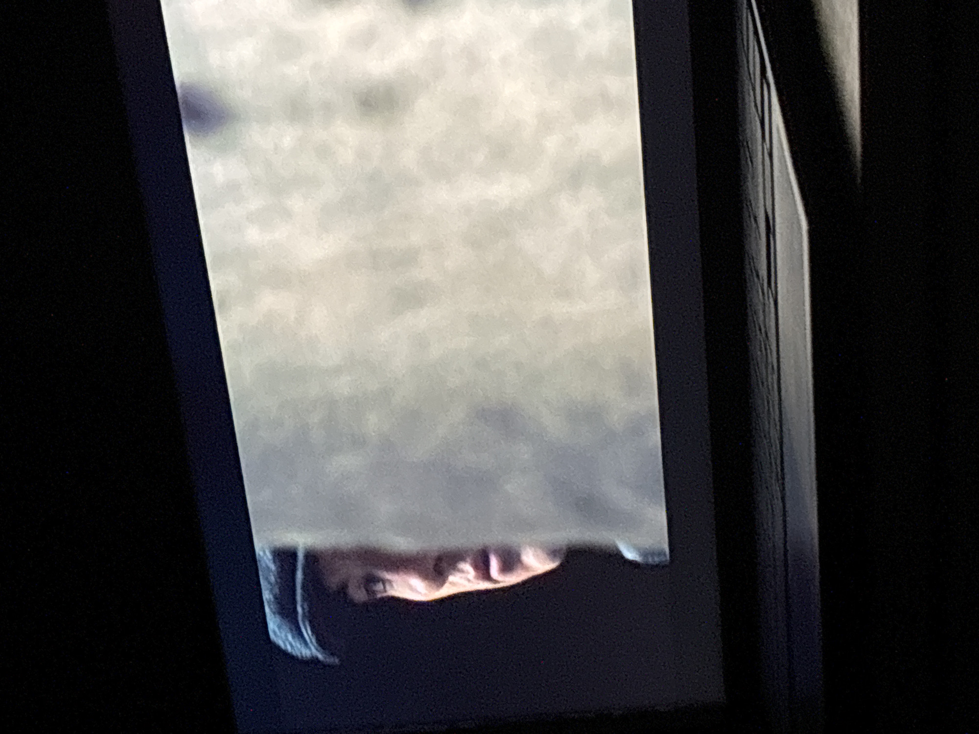 A screen displays what appears to be the lower portion of a person's face and part of their forehead, positioned at the bottom edge of the frame. The person has short hair, and the visible skin tone is fair. The upper part of the screen displays a blurry white/grey area. The screen is surrounded by a dark or black frame on three sides. The background surrounding the screen is dark. There is no identifiable information suggesting a location in Budva, Montenegro.