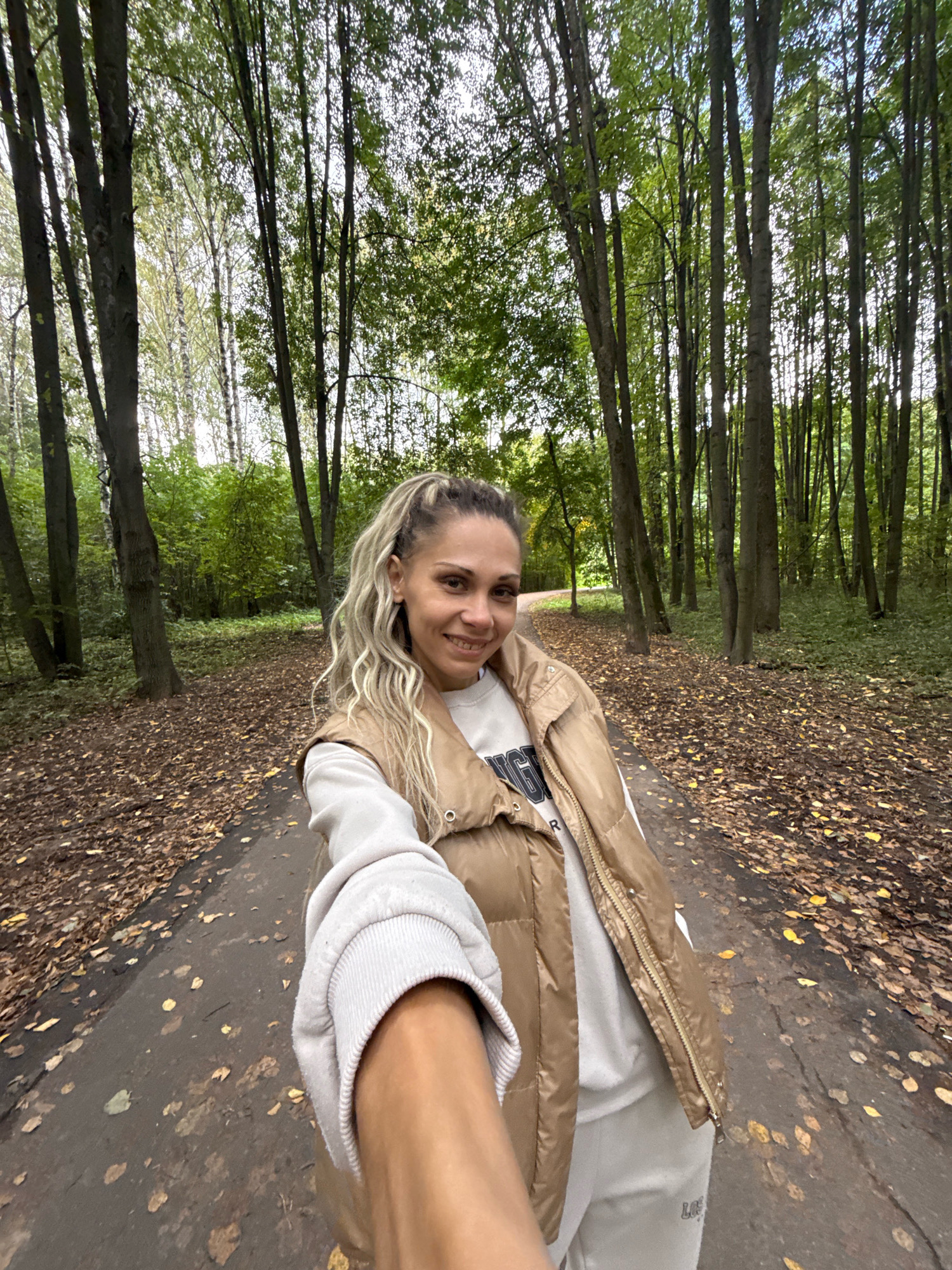 A young woman with long blonde hair tied in a ponytail, wearing a beige puffer vest over a white sweatshirt and light-colored sweatpants, smiles as she takes a selfie with her right arm extended. She is standing on a paved path in a forest area, likely in or near Rusanovo, Russia. The ground is covered with fallen brown and yellow leaves, indicating it is autumn. Tall trees with green and some yellowing foliage surround the path, with dappled sunlight filtering through the canopy. The path curves gently to the left in the background. The overall scene depicts a casual outdoor setting in a wooded environment.