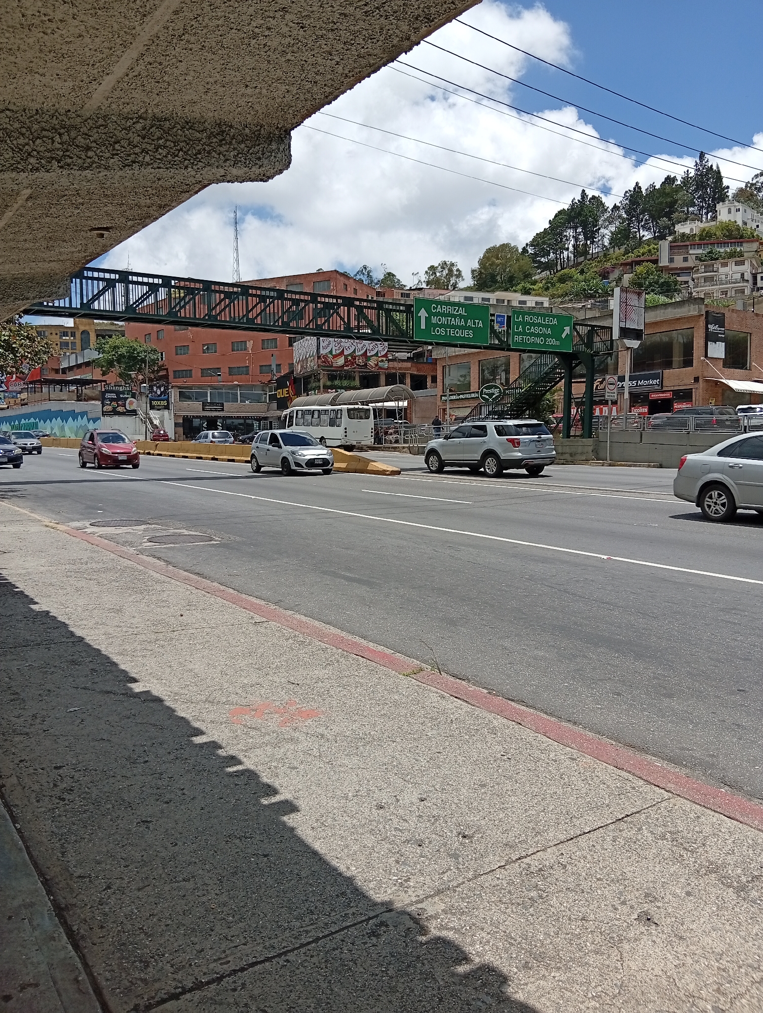 The image shows a street scene in Carrizal, Venezuela. A multi-lane road runs horizontally across the frame, with several cars driving on it. On the left side of the road, a pedestrian bridge spans across the lanes. Buildings are visible behind the bridge and along the side of the road, some with signs. A hillside covered in trees and houses rises in the background. The sky is blue with scattered white clouds. A shadow of an overhead structure falls across the sidewalk in the foreground.