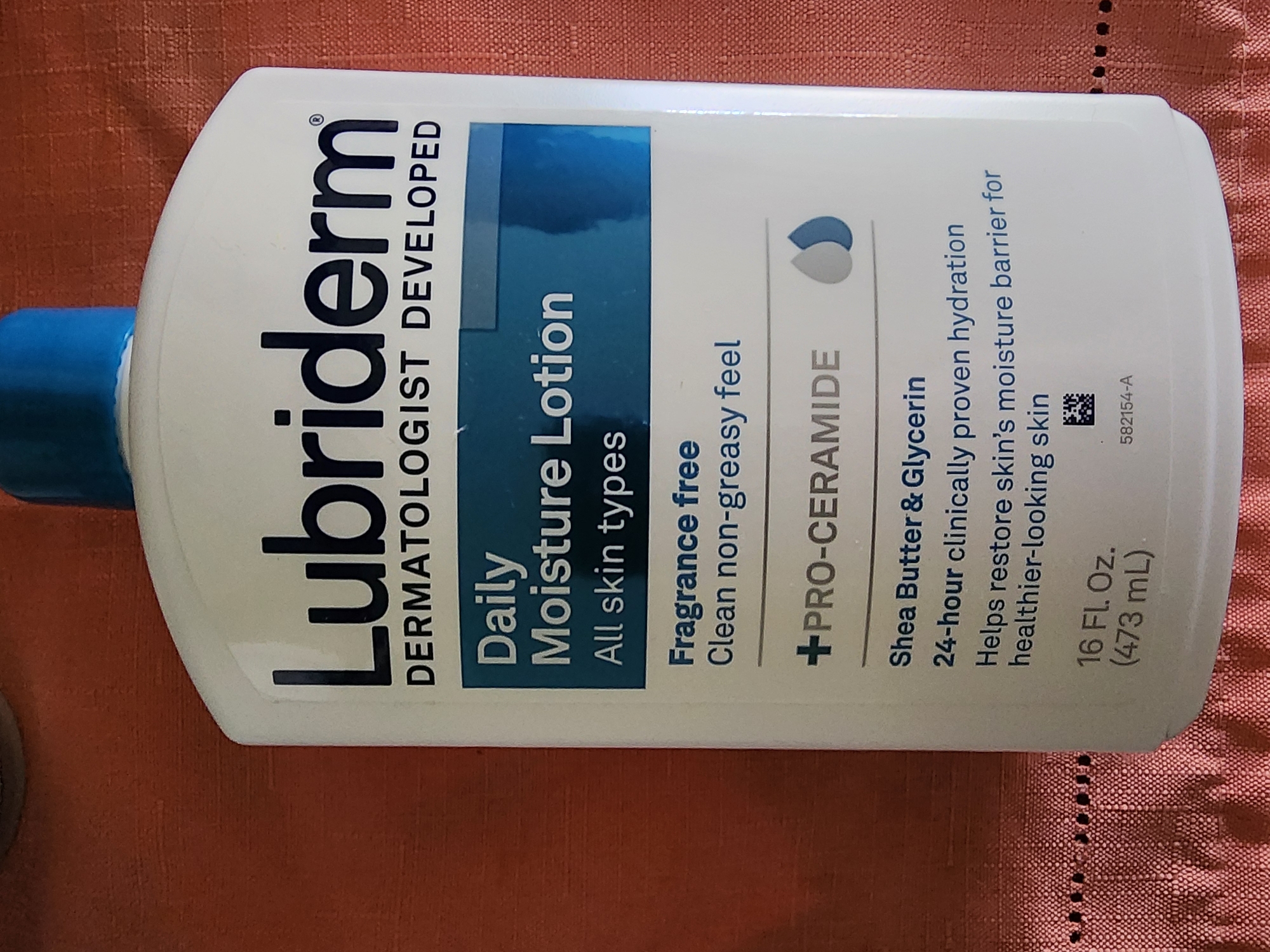 A white bottle of Lubriderm Daily Moisture Lotion is centered in the frame, positioned against a reddish-orange textured surface. The bottle has a blue cap and features black text for the brand name, product description, and key ingredients. The label indicates "DERMATOLOGIST DEVELOPED," "Daily Moisture Lotion," "All skin types," "Fragrance free," "Clean non-greasy feel," "+PRO-CERAMIDE," and "Shea Butter & Glycerin." Below that, it states "24-hour clinically proven hydration Helps restore skin's moisture barrier for healthier-looking skin." The volume is listed as "16 FL. Oz. (473 mL)". A small barcode is visible on the lower right side of the bottle. The environment appears to be indoors, with the textured surface potentially being a tablecloth or fabric. There are no people or actions depicted. The image is a close-up of the product, suggesting it is for informational or product identification purposes. The specific location within Lower Paxton Township, United States of America, is not visually discernible from the provided image.