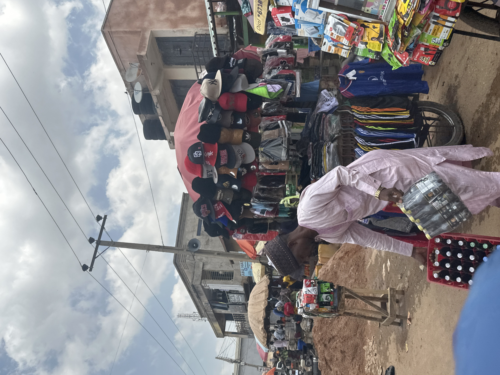 The image depicts a bustling outdoor market scene, likely in Jos, Nigeria. The viewpoint is from a low angle, looking upwards towards a cloudy sky and some buildings. In the foreground, a person dressed in traditional light pink attire is partially visible, holding a pack of bottled drinks. To their right, a red crate overflowing with dark-bottled beverages is also prominent.
The market stalls are filled with various merchandise, including a large display of baseball caps of different colors and designs, neatly stacked on a red umbrella-shaped awning. Behind the caps, more items are visible, such as clothing and accessories like electronics chargers, indicated by the visible text "9D9Tech" and "STRONG FAST CABLE." Other text fragments like "ABU HAS" and a phone number "#671#" are also discernible on signs.
The overall impression is one of a vibrant, active marketplace with goods being displayed and sold. The time of day appears to be daytime, given the lighting, and the sky suggests it might be a slightly overcast or cloudy day. The presence of the market stalls, the variety of goods, and the traditional attire hint at a local cultural setting. The ground is uneven and appears to be dusty or dirt-covered, common in such outdoor market environments. The scene is lively and suggests commercial activity is in full swing.