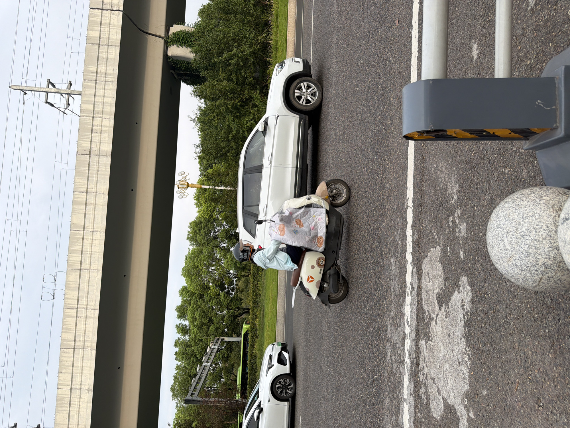 In Jiangjin, China, a person wearing a light blue jacket and a black helmet is riding a scooter. The scooter is light-colored with some red and brown accents and has a decorative covering on the front, featuring cartoon characters. The scooter is on an asphalt road, with a white line marking the lane. A white SUV is to the right of the scooter, also on the road. A white car is visible to the left, partially cut off by the frame. In the background, trees and a large concrete overpass are present. The sky is overcast.