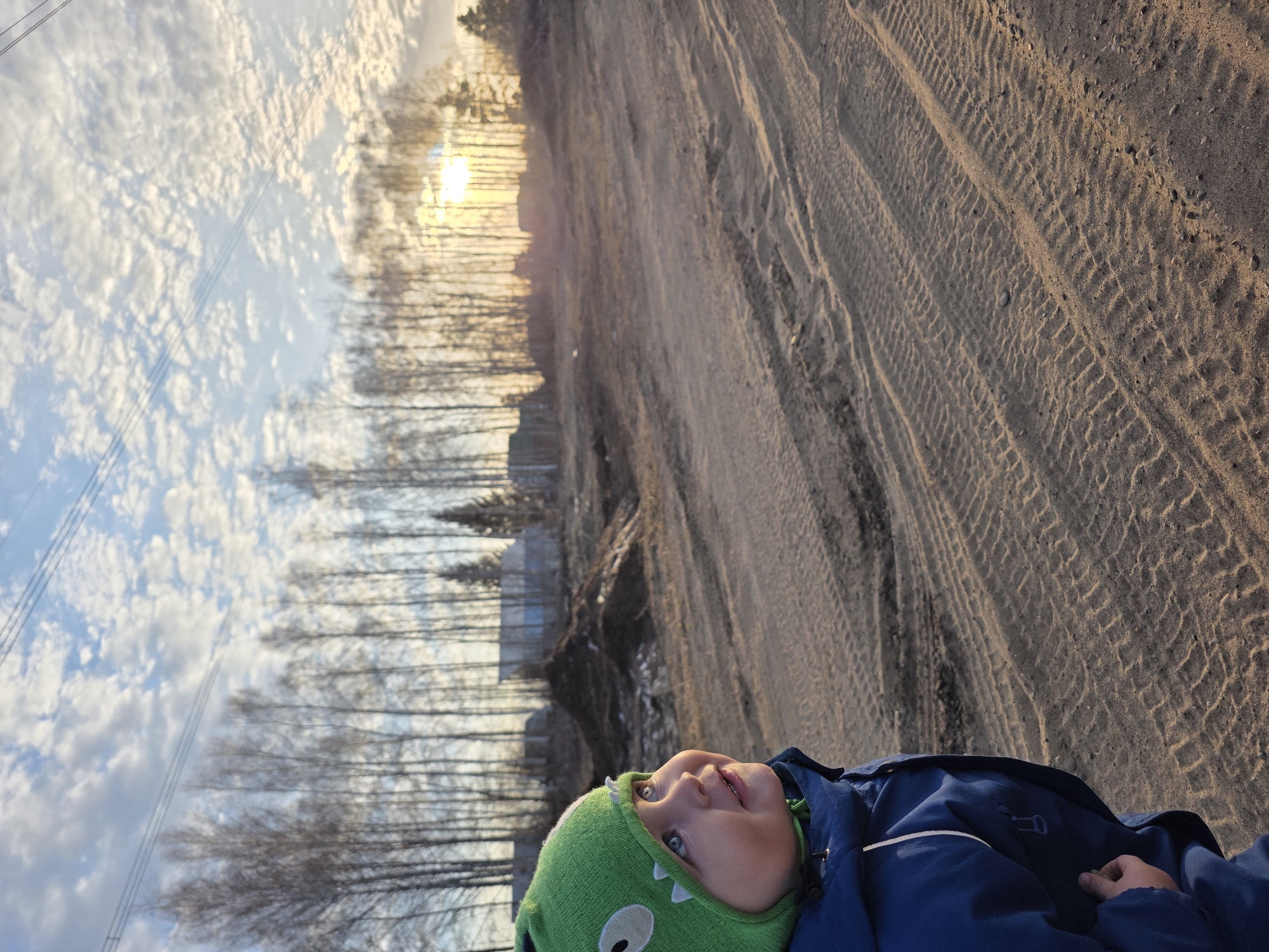 A child wearing a green hat and a blue jacket lies on the ground. The ground is sandy with tire tracks. In the background, there is a line of trees and a sky with clouds and the sun. The location is Noviy, Russia.