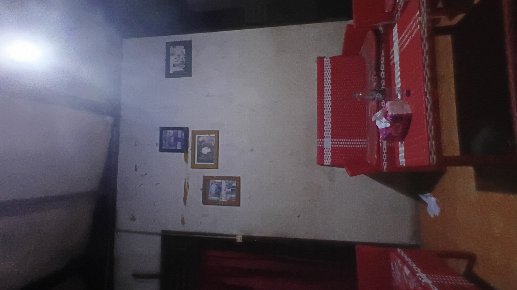The image depicts an indoor scene, likely a living room or common area. A red woven bench is placed in the foreground, topped with a tissue box and a plastic bottle of water. To the left, another red woven chair is partially visible. The walls are a light, textured color, adorned with three framed photographs. The ceiling is visible at the top, with what appears to be exposed beams and a bright light source illuminating the space. The floor is a warm, orange-brown color. The overall impression is of a simply furnished domestic interior. There is no indication of the specific location within Prabumulih, Indonesia, or any human presence within the frame.