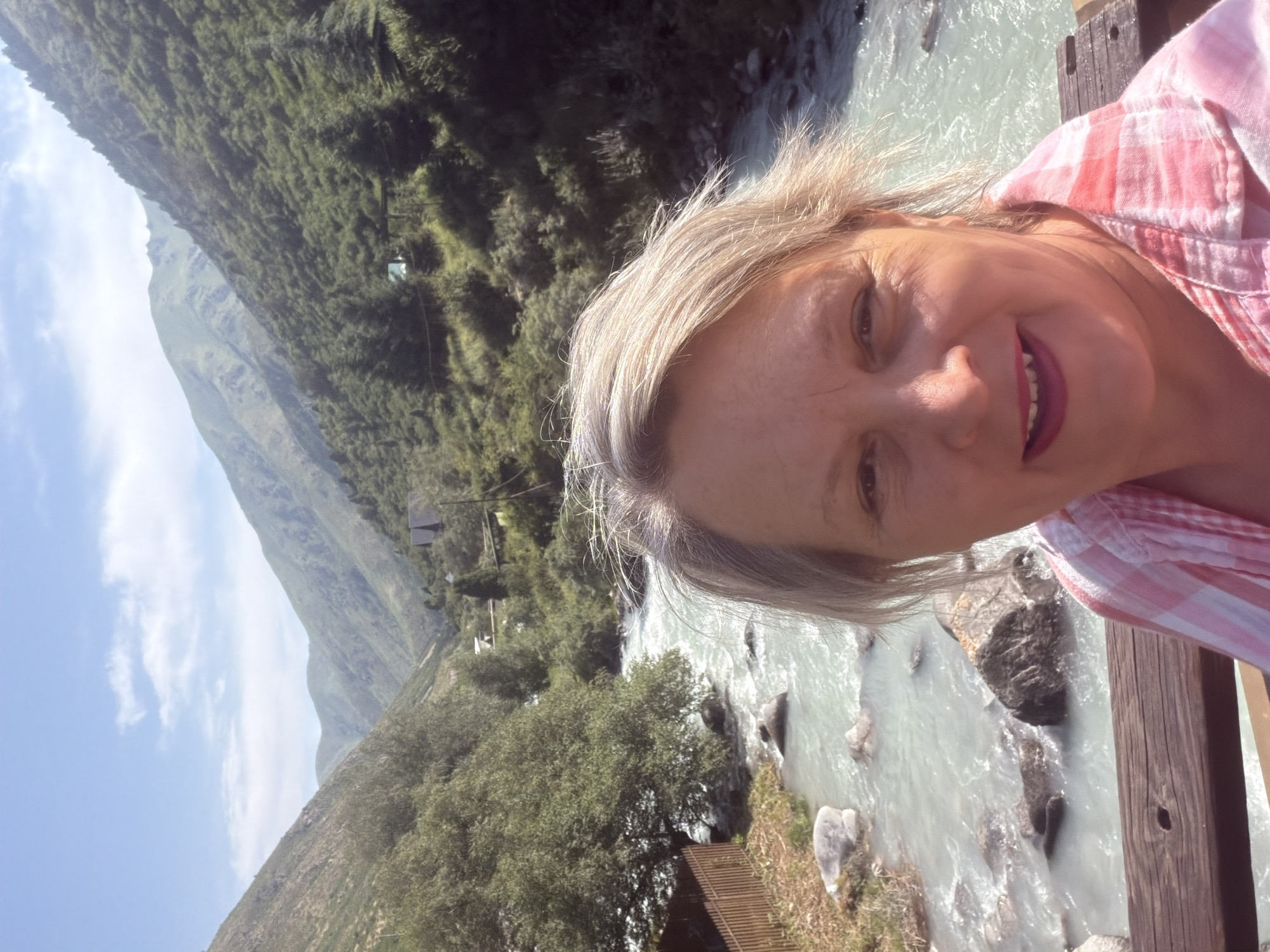 A woman with blonde hair and bright pink lipstick smiles at the camera in a selfie taken outdoors. She is wearing a pink and white checkered shirt. Behind her, a fast-flowing river with rocks is visible. The river is surrounded by lush green trees and steep, mountainous terrain. The sky is blue with a few white clouds. The location is a natural landscape, possibly a mountain valley with a river.