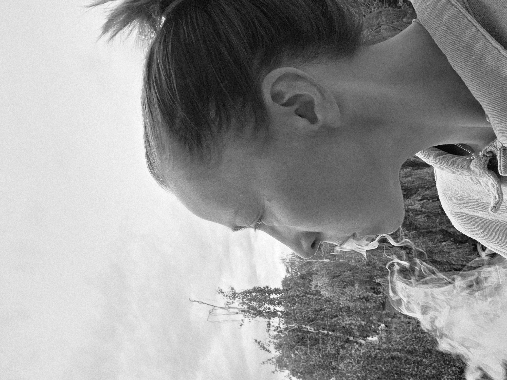 A black and white close-up image depicts a person with their head tilted downwards, appearing to exhale smoke. The person has their hair pulled back in a bun. Their profile is visible, and a portion of a jacket is visible on their right shoulder. In the background, there is a blurred image of trees. The location and city of Chaikovsky, Russia cannot be determined from this image.